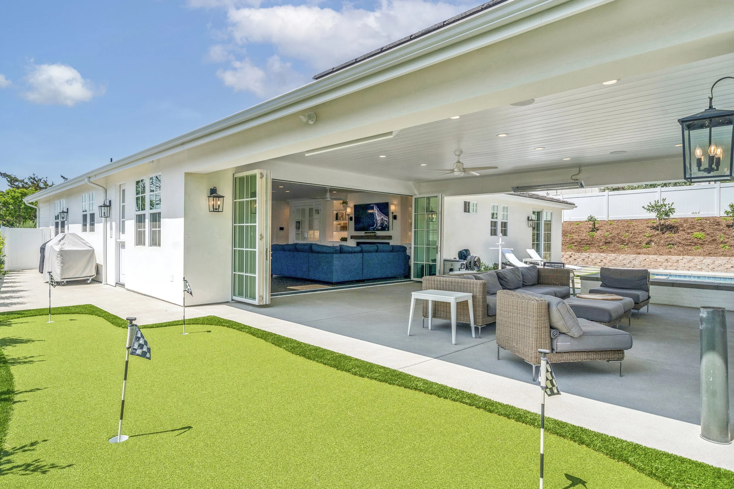 Covered outdoor living area with lounge seating, folding glass doors, and a backyard putting green at a remodeled Orange County home.