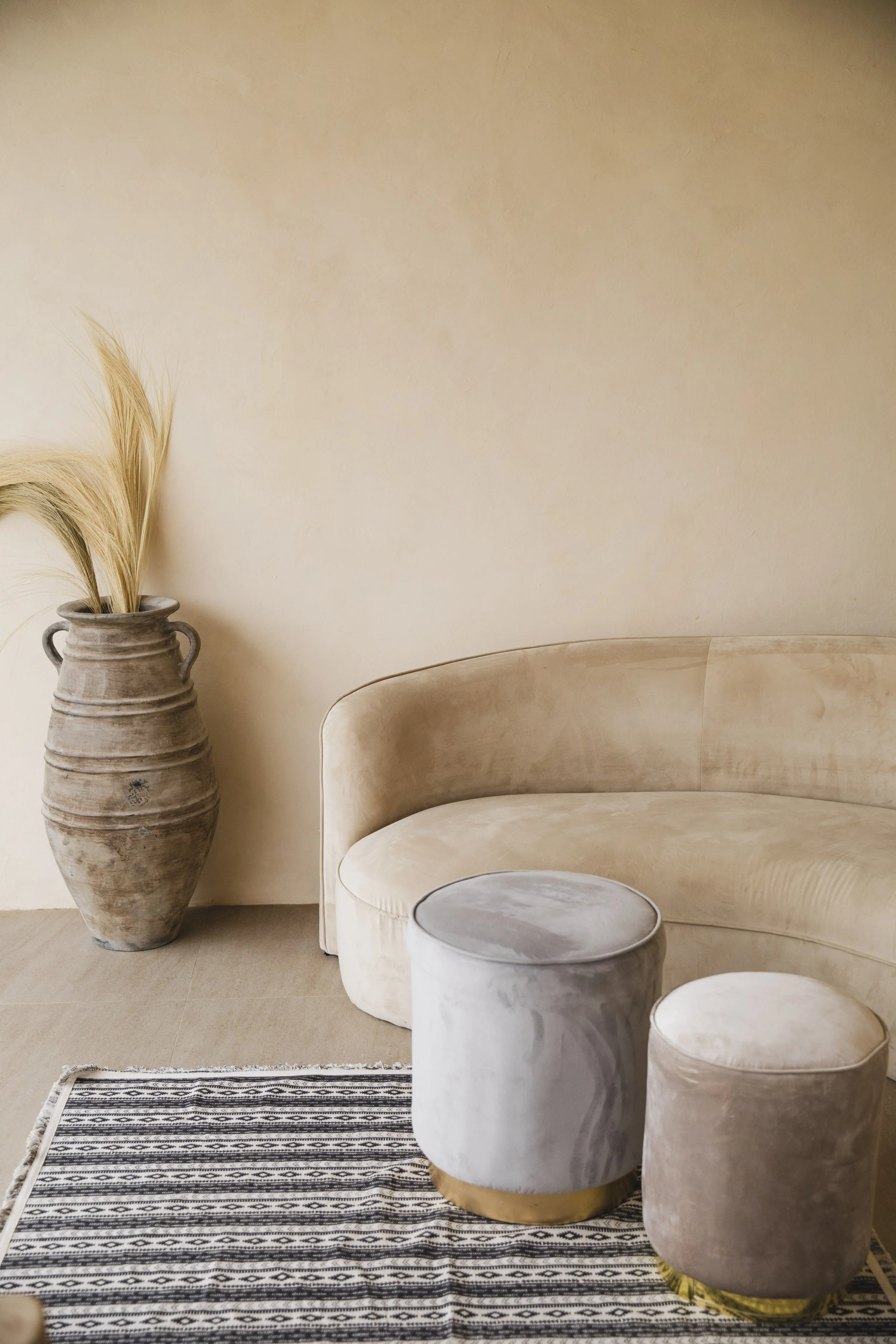 Minimalist living room with a curved neutral sofa, velvet poufs, vintage ceramic floor vase, textured rug, and warm organic finishes.