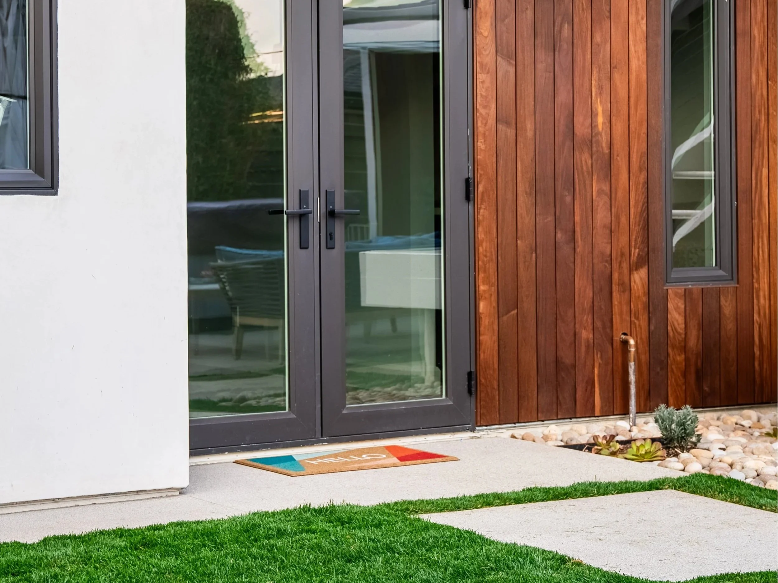 Close-up of the ADU entrance featuring black framed glass doors, wood siding detail, and concrete walkway surrounded by artificial turf and landscaping.
