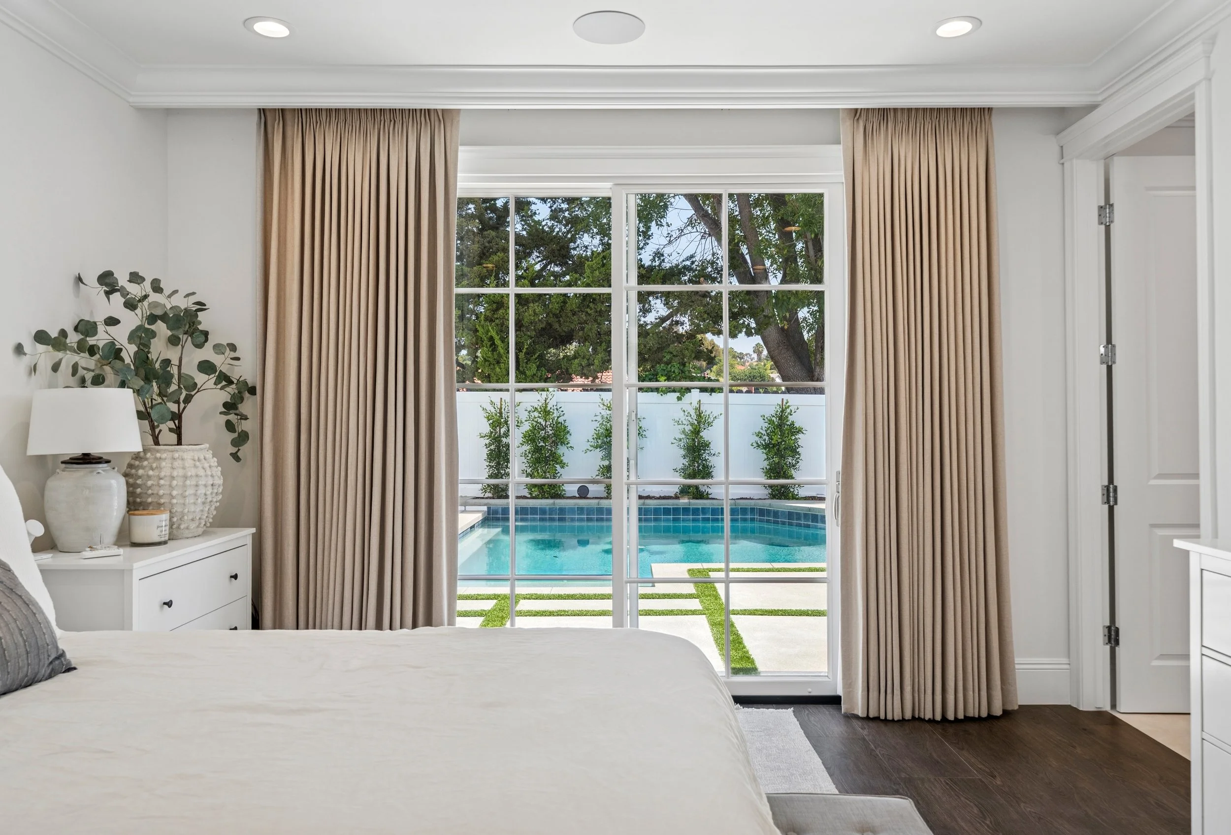 Primary bedroom with large glass doors, neutral drapery, white walls, wood flooring, and views of the backyard pool in a Mission Viejo custom home.