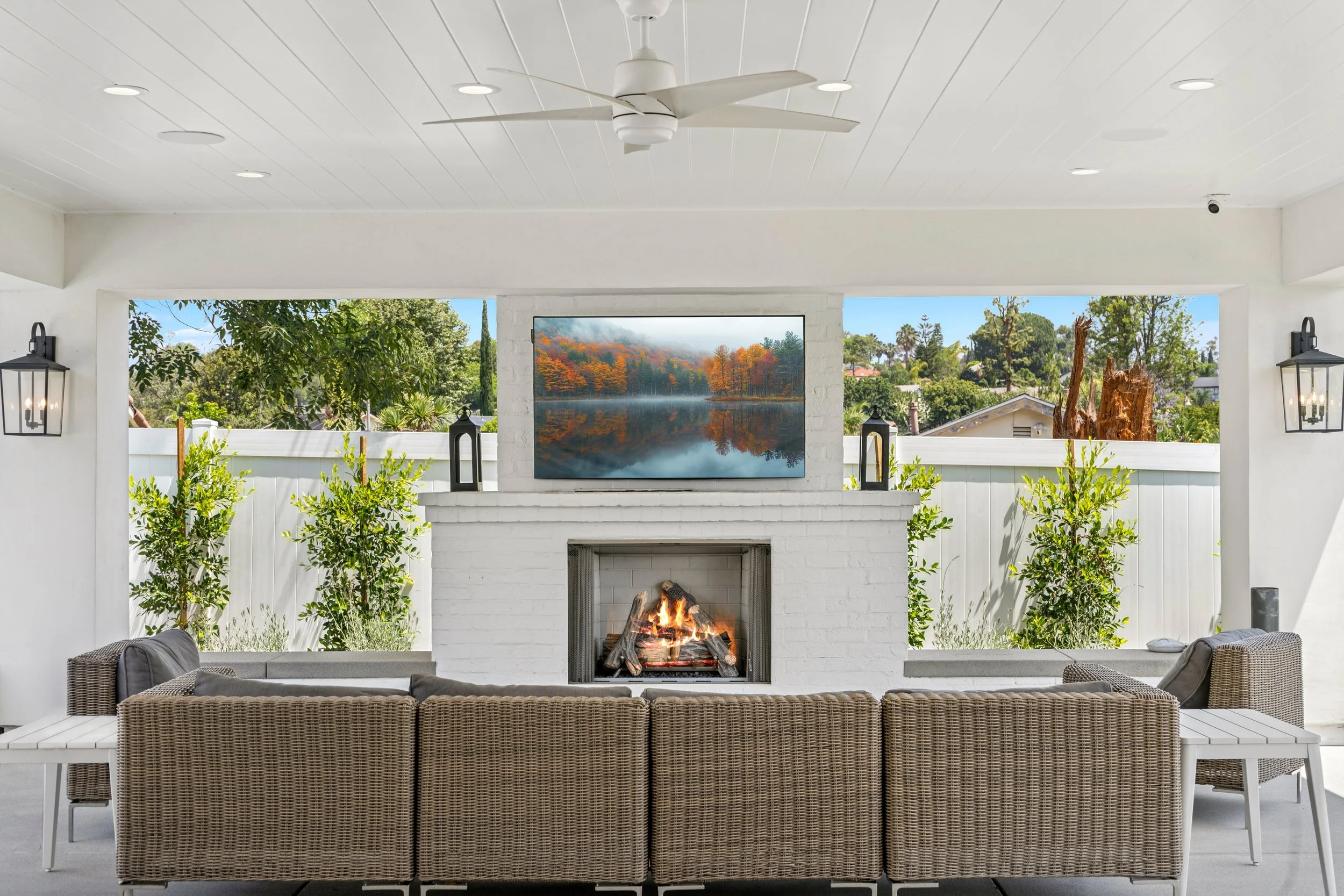 Covered outdoor living area with white brick fireplace, mounted TV, wicker lounge seating, ceiling fan, and backyard greenery.