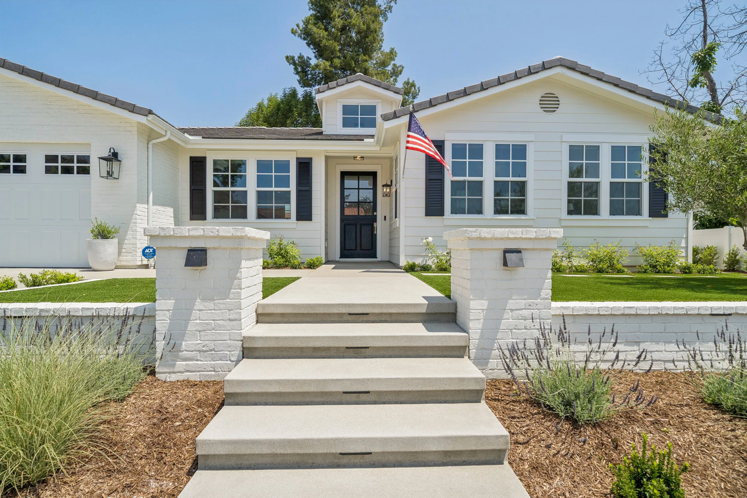 Front exterior of a renovated white coastal farmhouse-style home with fresh landscaping, white brick, black shutters, and curb appeal in Orange County.