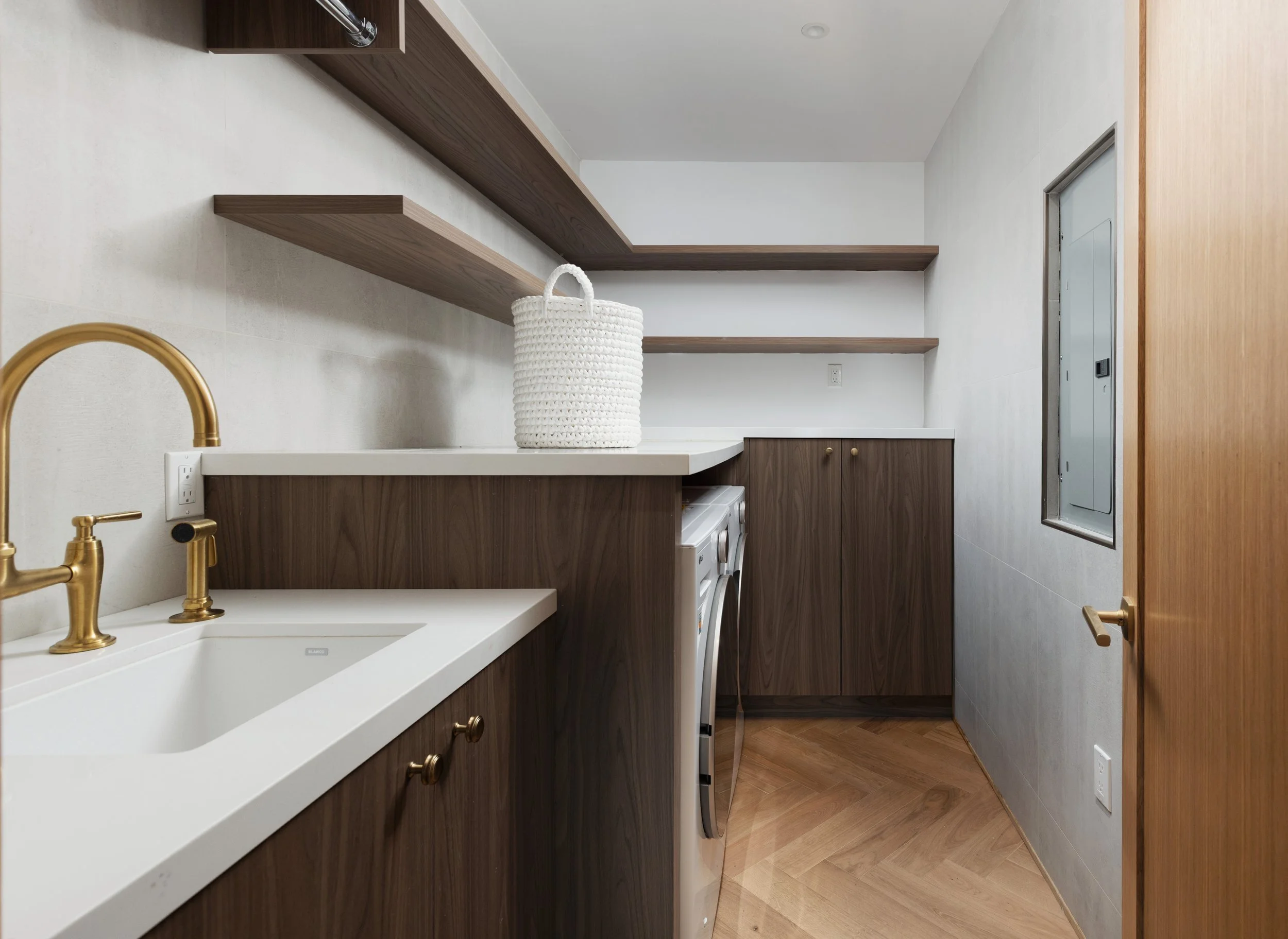Modern laundry room with walnut cabinetry, brass faucet, white utility sink, open shelving, and herringbone wood flooring.