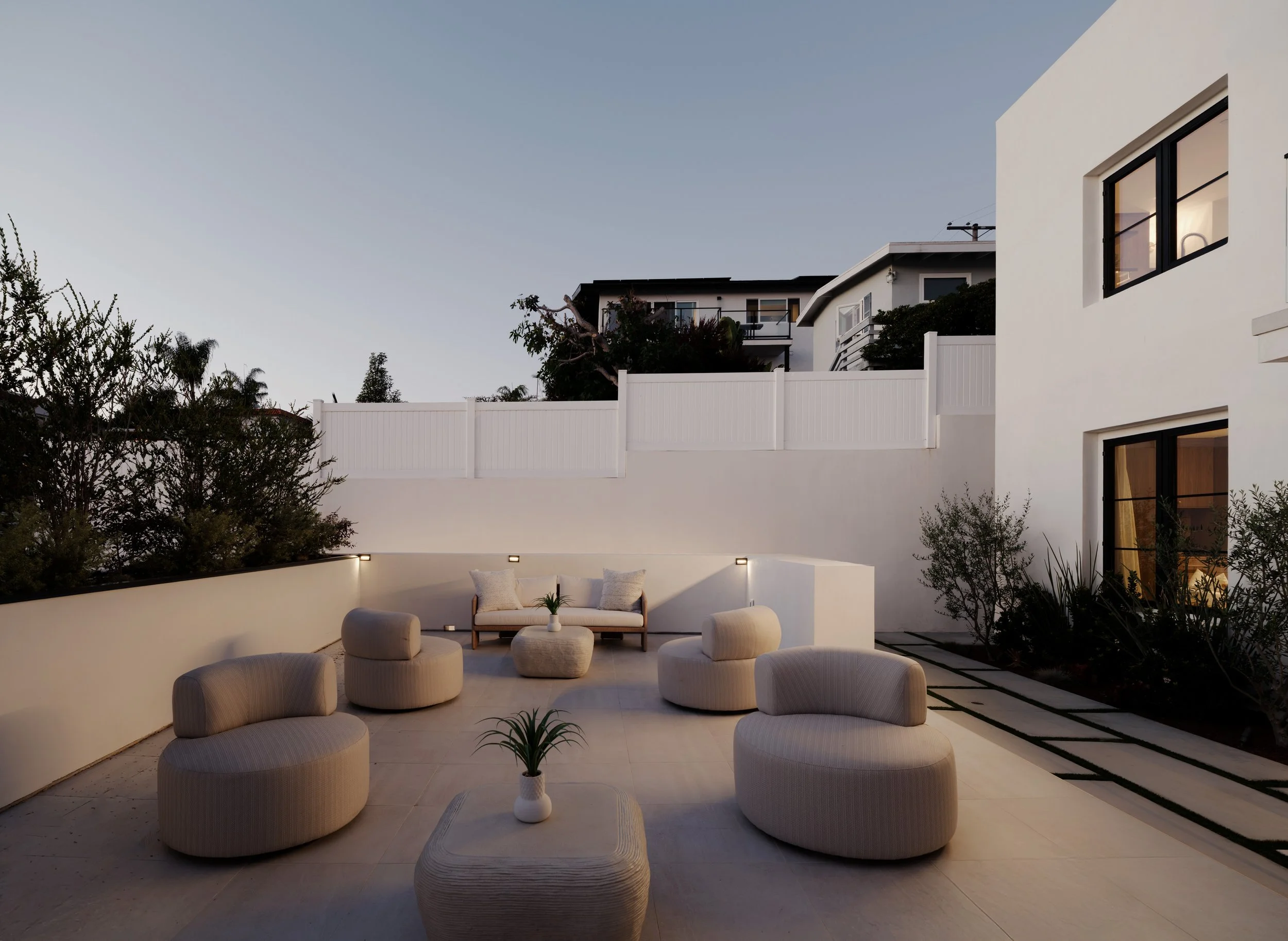 A modern, minimalist outdoor patio at dusk featuring cream-colored rounded swivel chairs and a matching sofa arranged around low stone coffee tables.