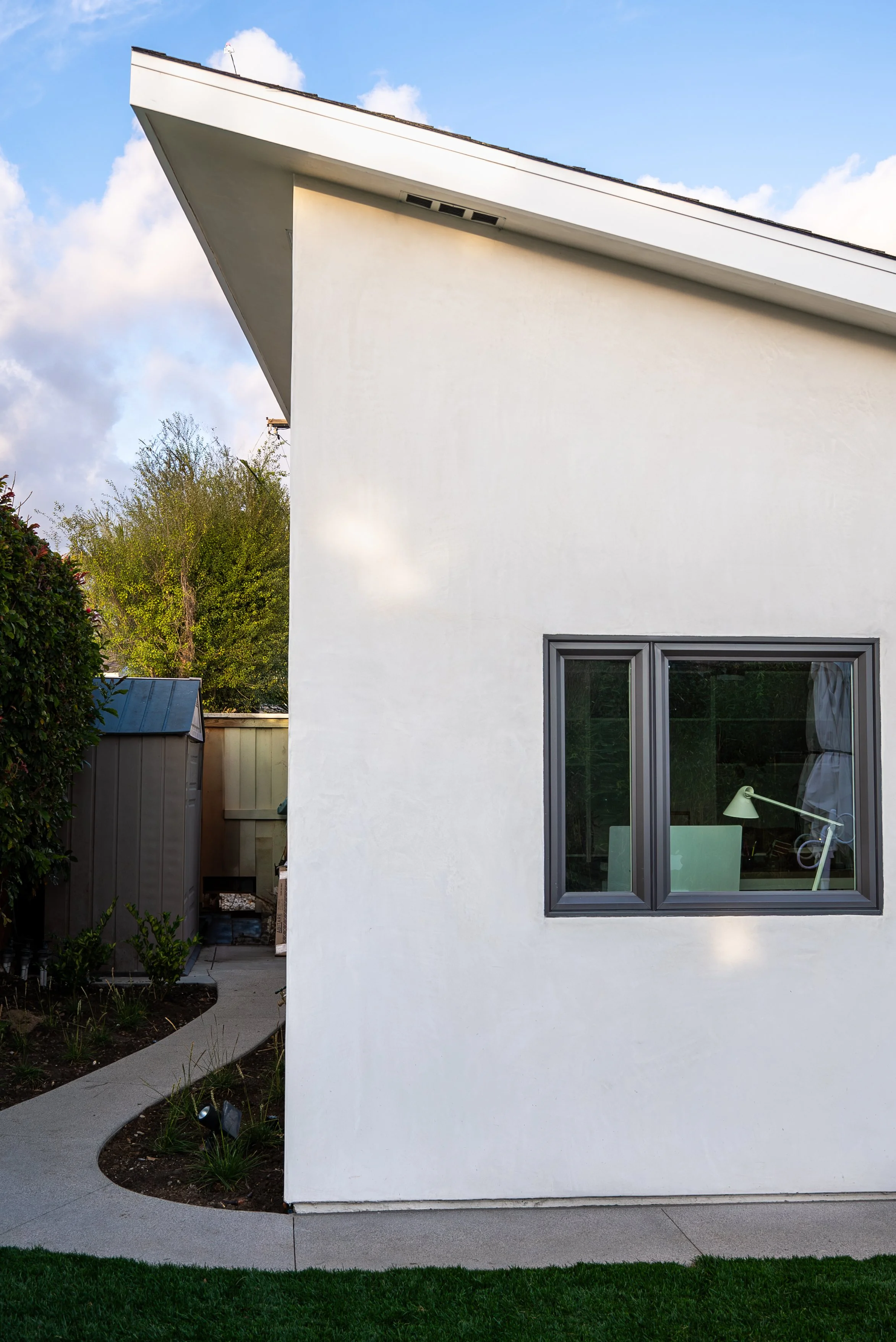 Side exterior view of a modern ADU with smooth white stucco, gray-framed window, sloped roofline, curved walkway, and surrounding landscaping.