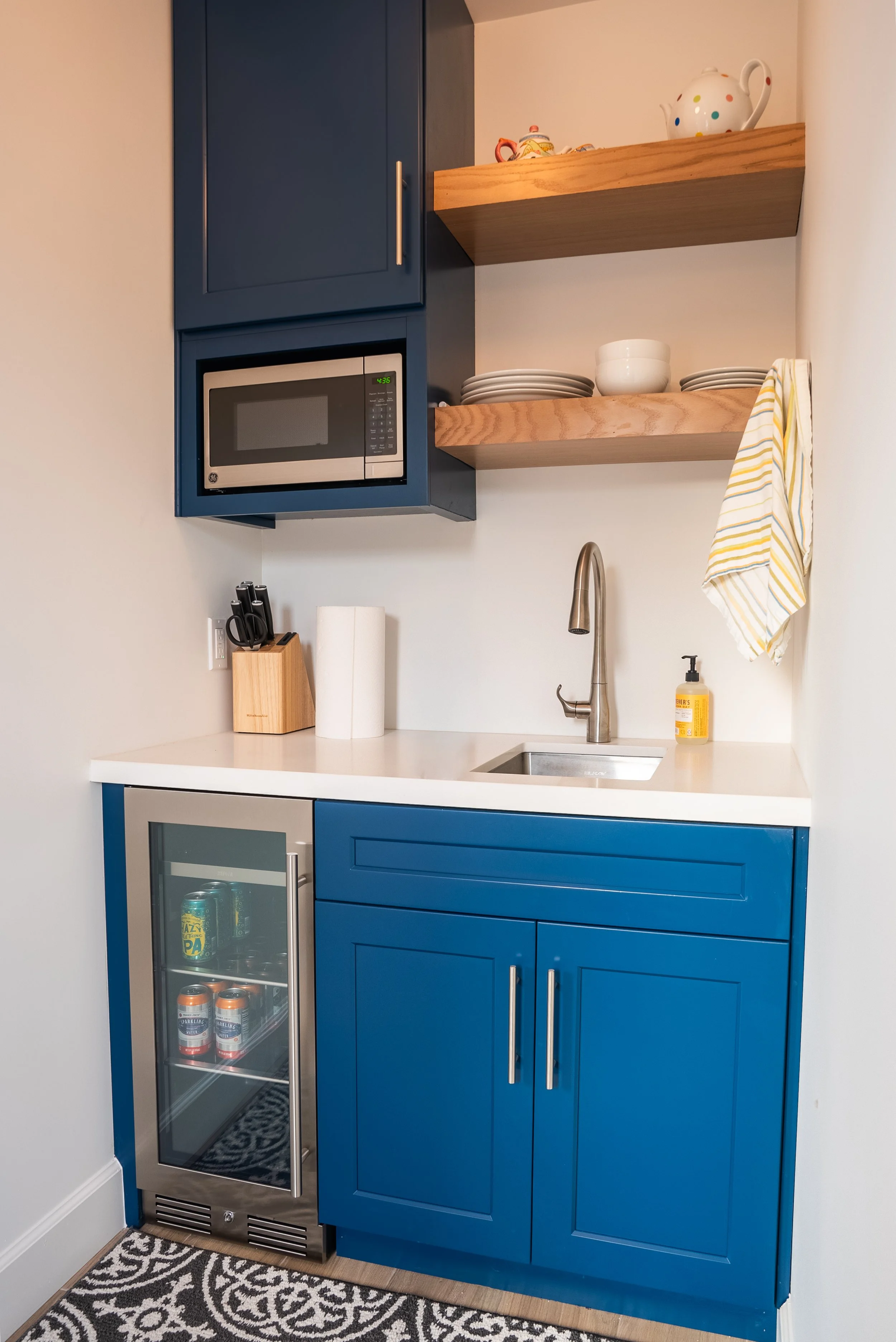 Compact ADU kitchenette with blue cabinetry, white countertop, built-in microwave, beverage fridge, sink, and open wood shelving.