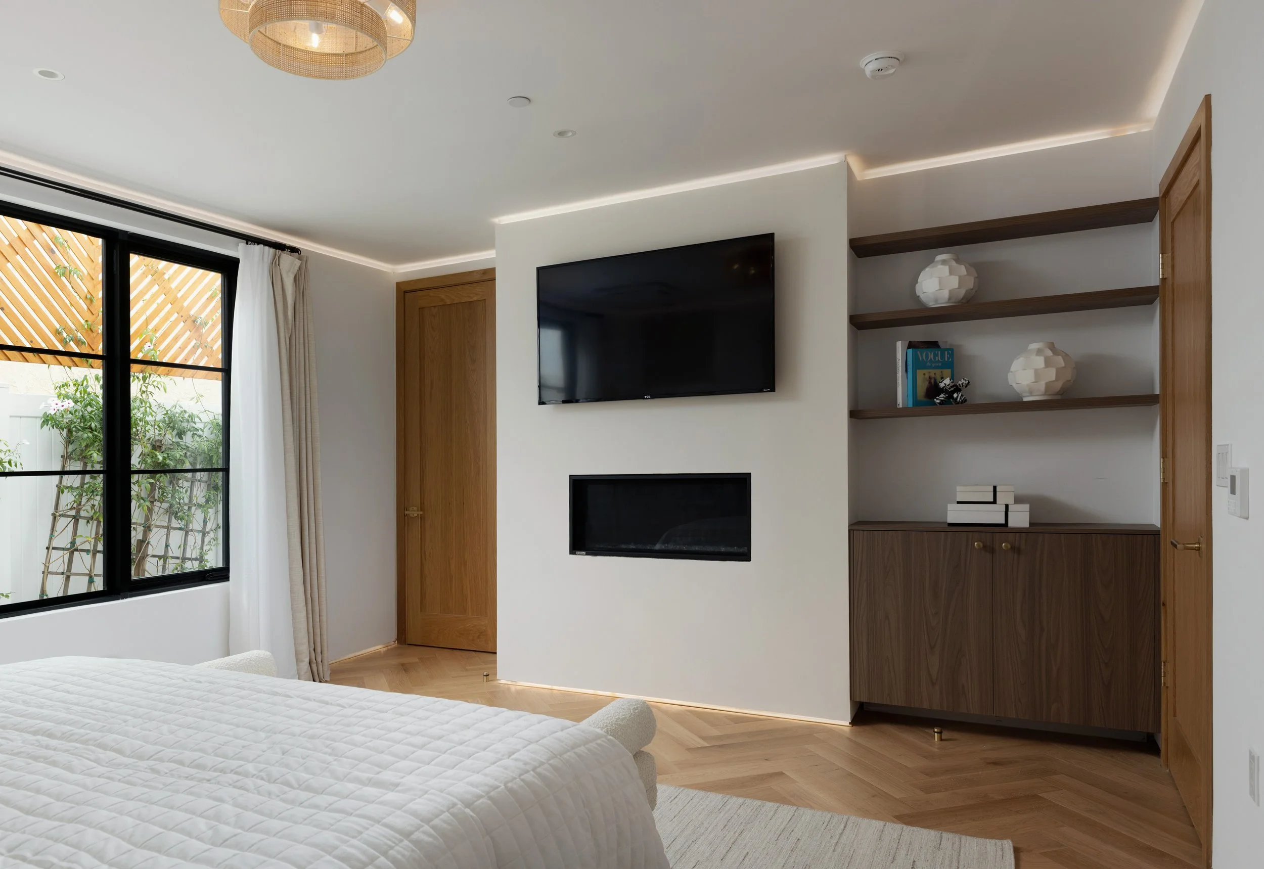 Primary bedroom with wall-mounted TV, modern fireplace, walnut built-ins, black framed windows, and warm wood flooring.