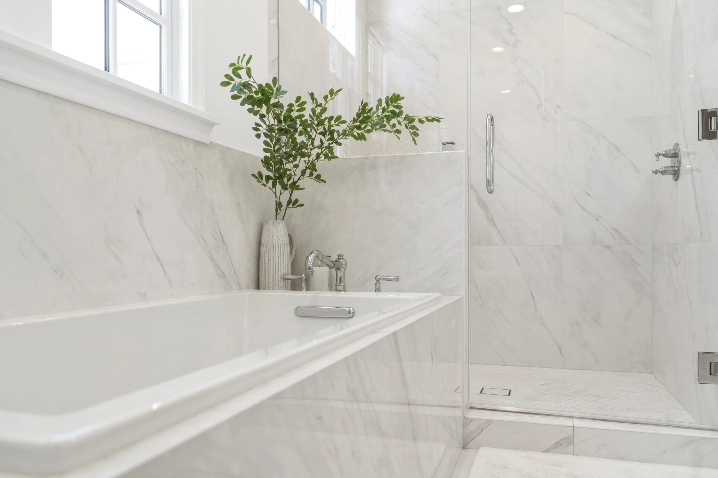 Close-up of a primary bathroom with marble shower, freestanding tub, chrome fixtures, glass shower enclosure, and soft natural light.