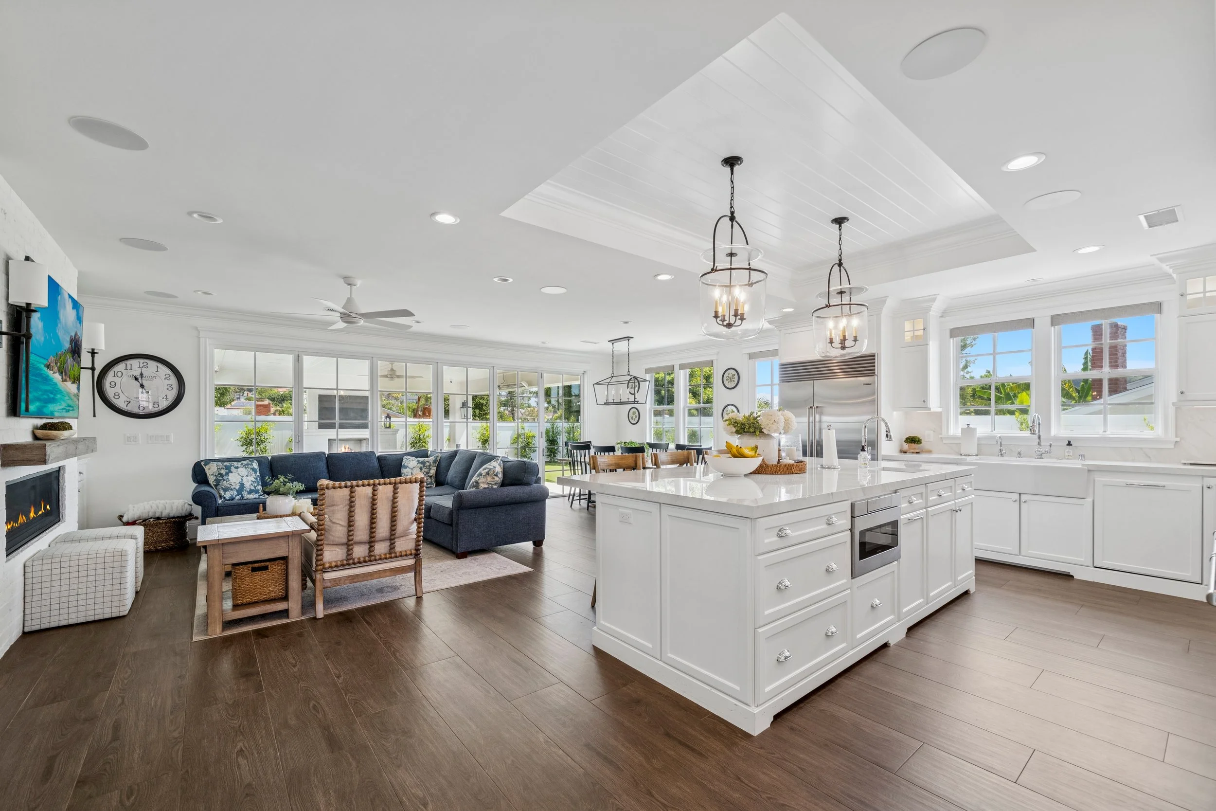 Bright custom kitchen and living space with white shaker cabinets, large island, farmhouse sink, stainless refrigerator, wood flooring, and open-concept layout.