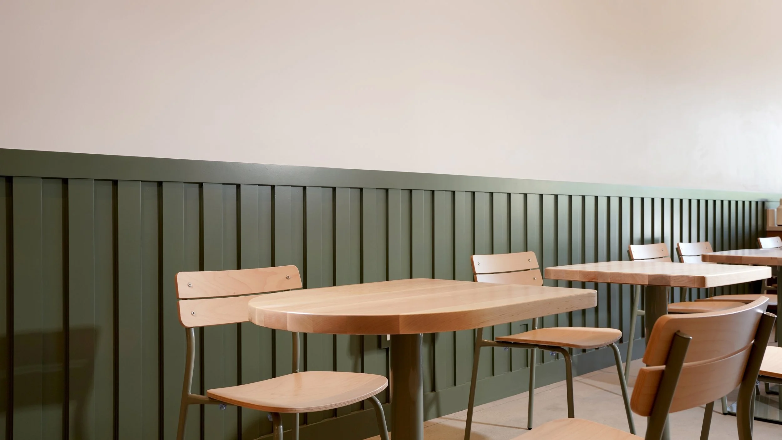 Coffee shop dining area with light wood tables, minimalist chairs, green vertical paneling, and clean commercial interior finishes.