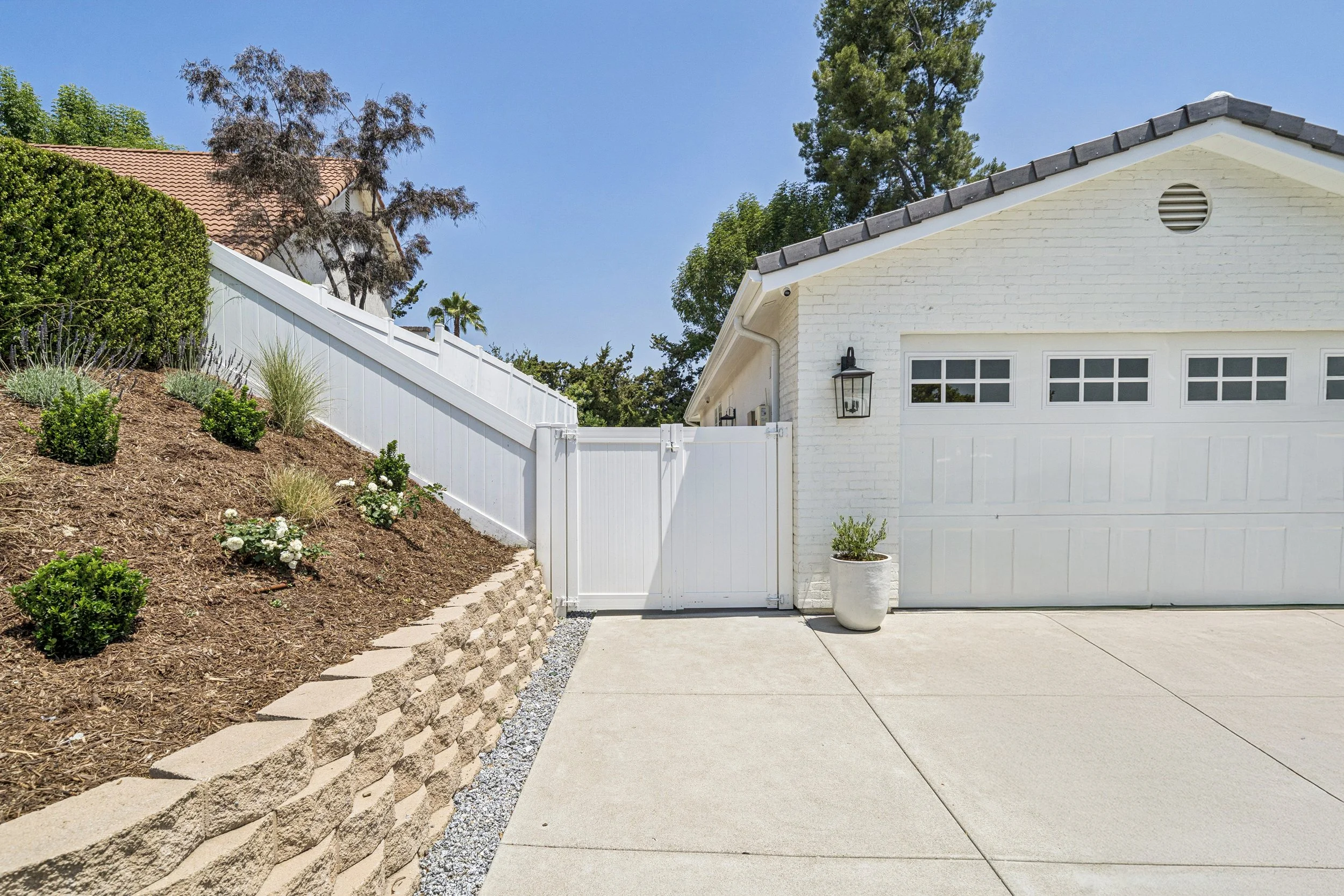 Side driveway view showing a white painted brick garage, white privacy gate, retaining wall, and refreshed hillside landscaping.