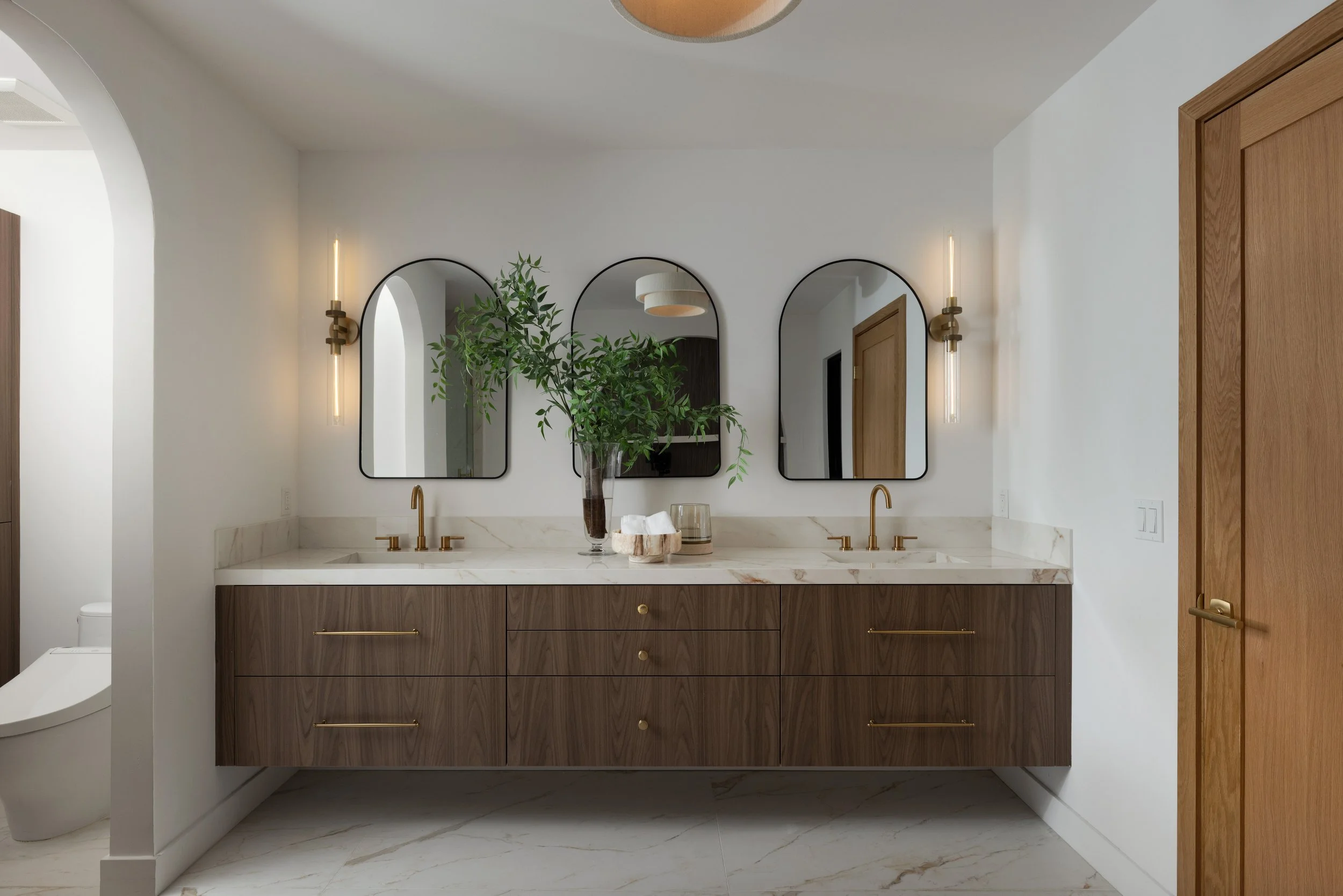 Modern bathroom vanity with walnut cabinets, brass fixtures, arched doorway, marble-look counters, and organic modern styling.