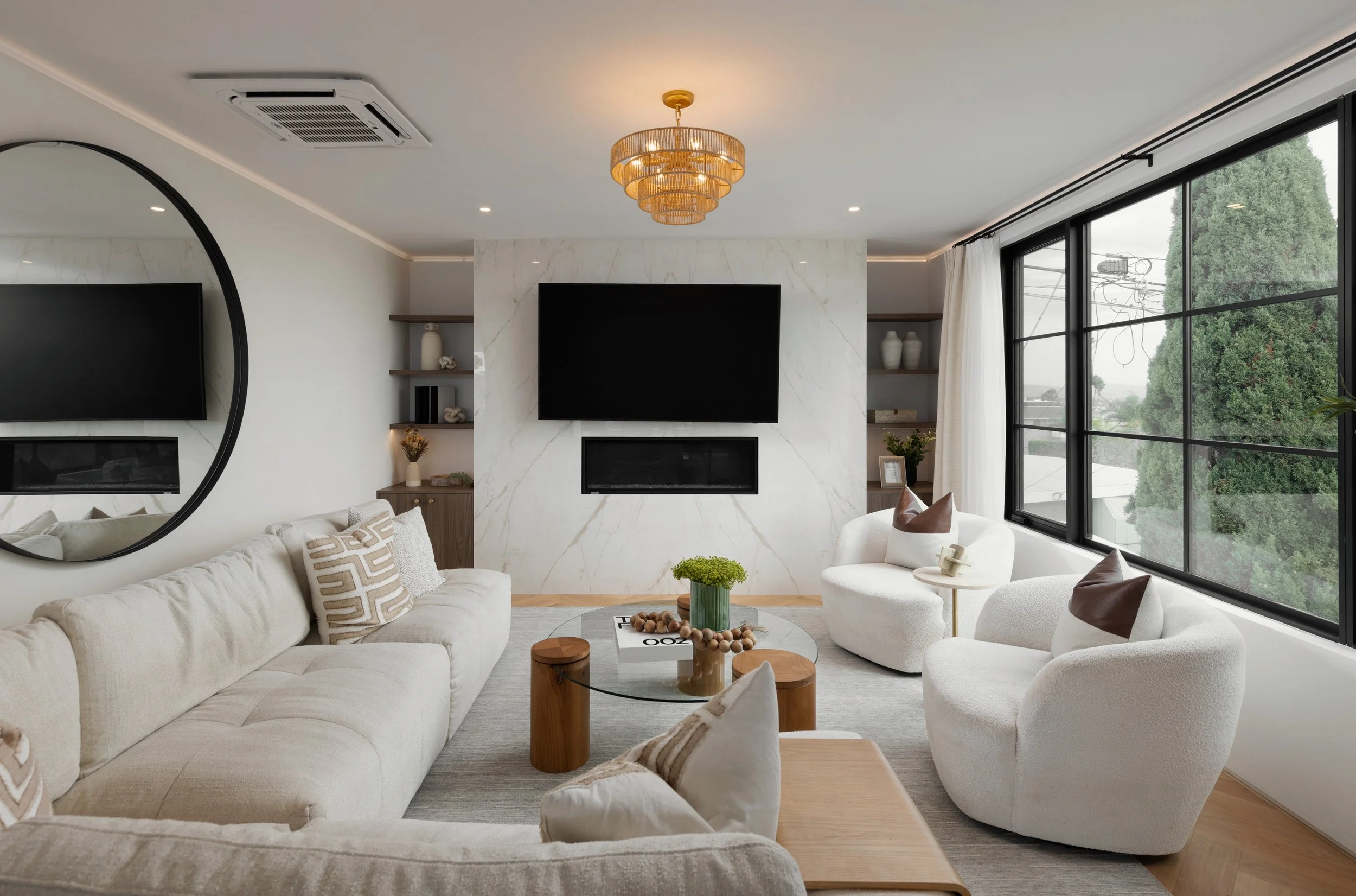 Coastal modern living room with cream chairs, wood coffee table, large abstract art, black windows, and natural greenery.
