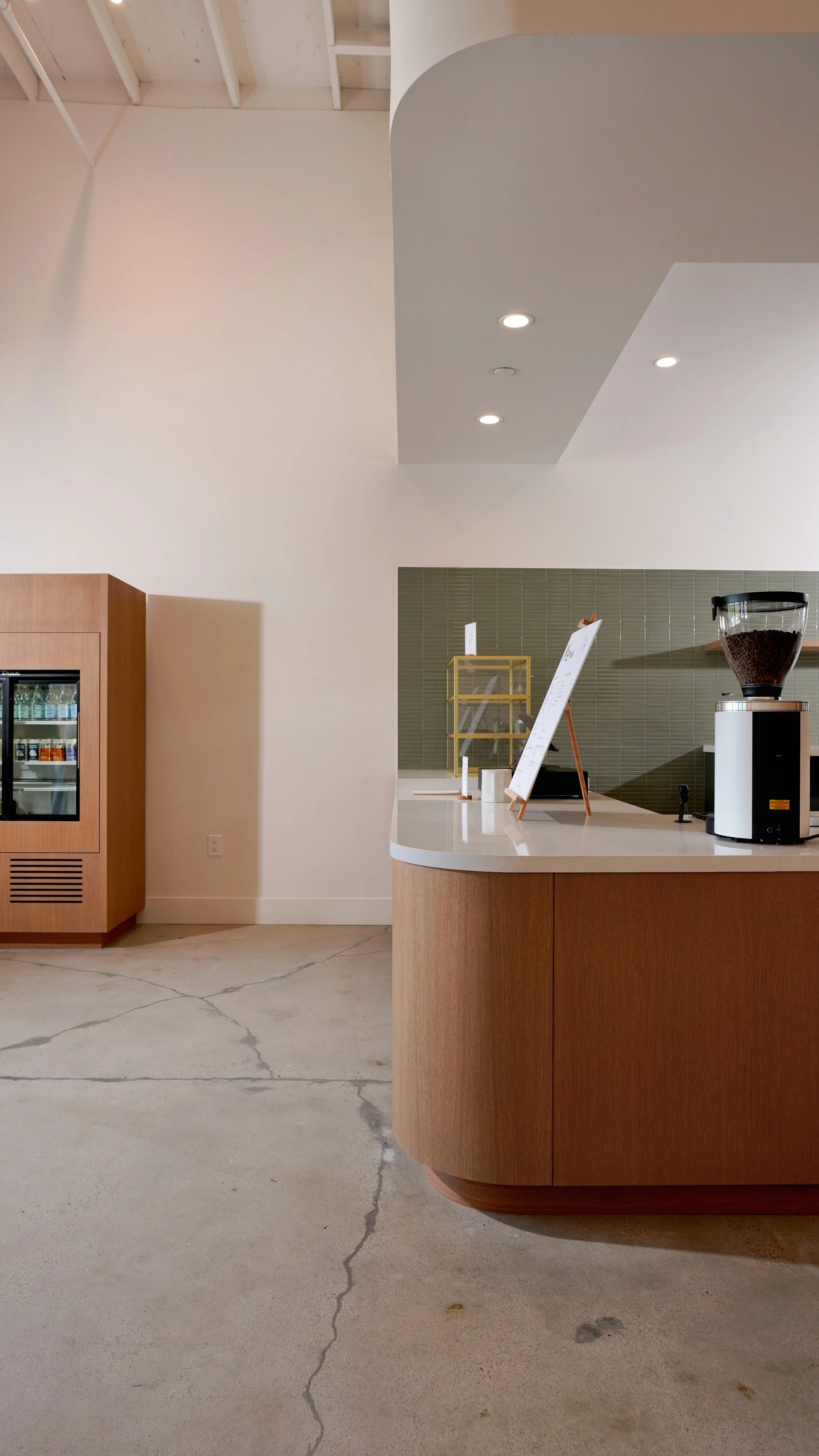 Vertical view of a coffee shop service counter with wood cabinetry, white countertop, green tile wall, and polished concrete flooring.