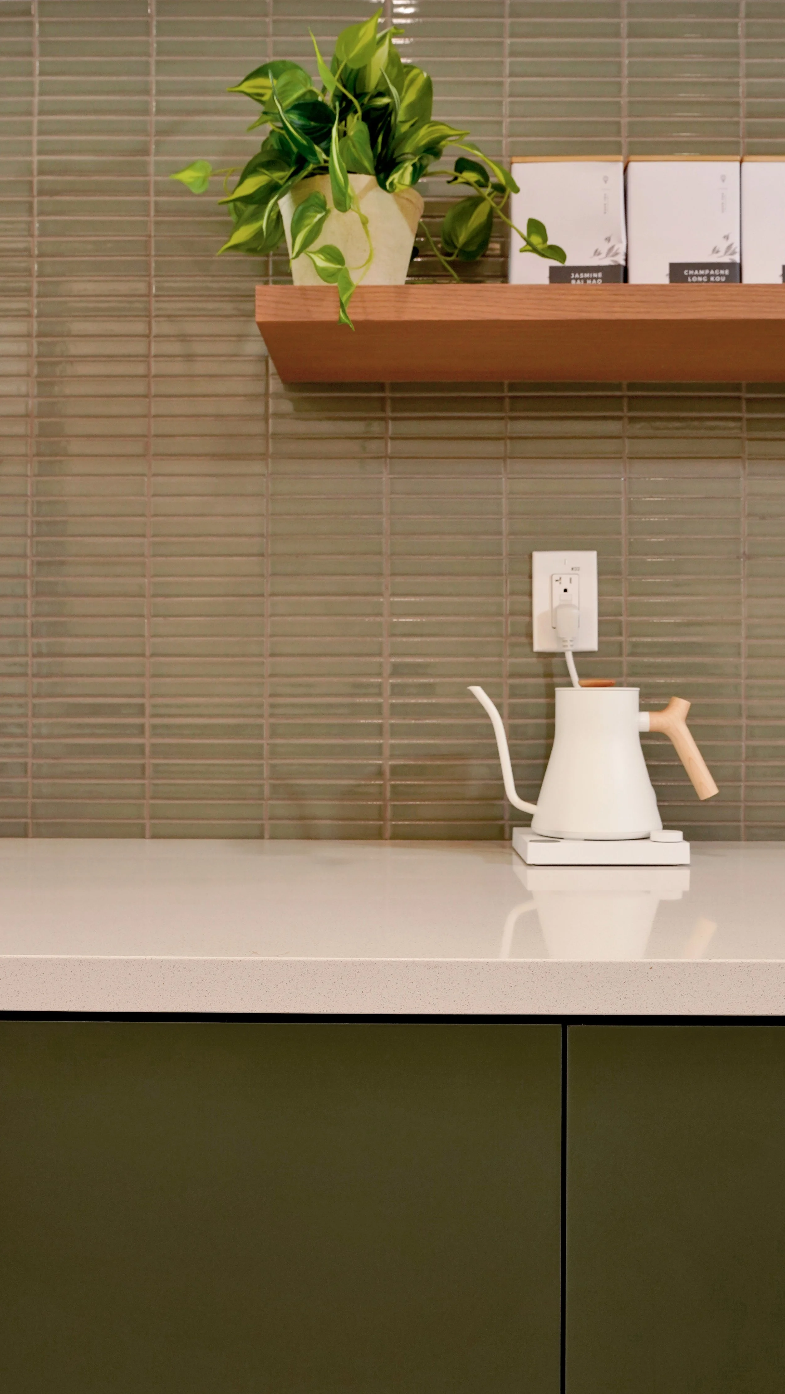 Coffee shop counter detail with green lower cabinetry, brass faucet, white countertop, stacked tile backsplash, and open wood shelving.