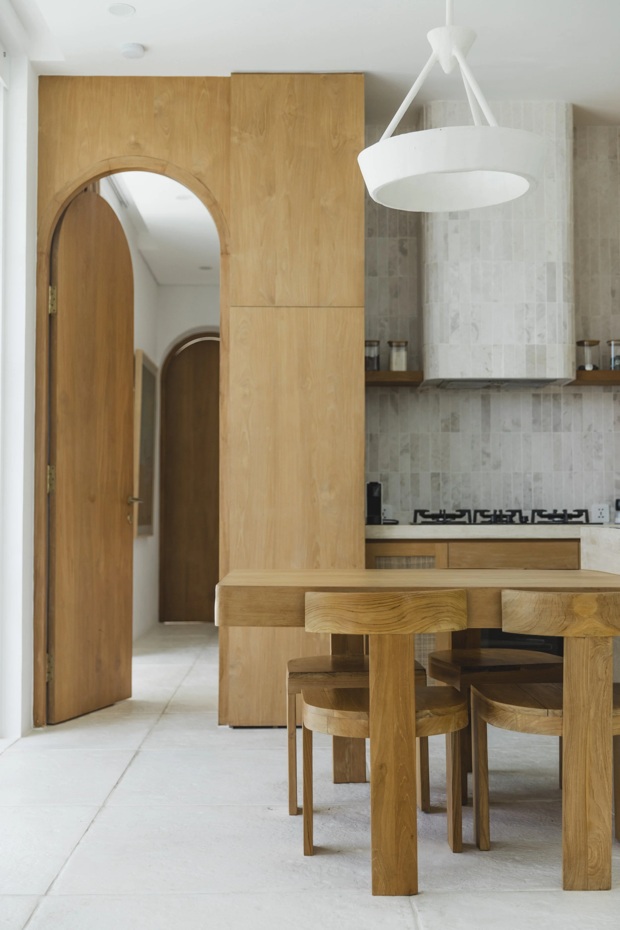Modern kitchen and dining area with natural wood cabinetry, arched doorways, stone tile surfaces, and a sculptural pendant light.