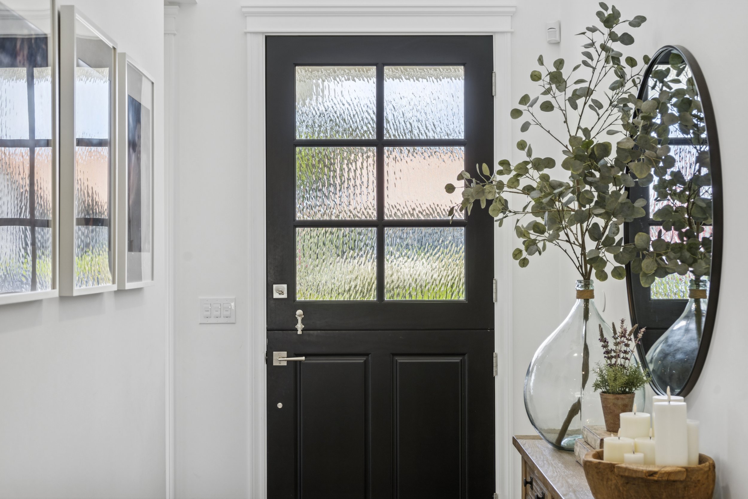 Entryway detail with black front door, white walls, framed art, round mirror, wood console, candles, and natural greenery.