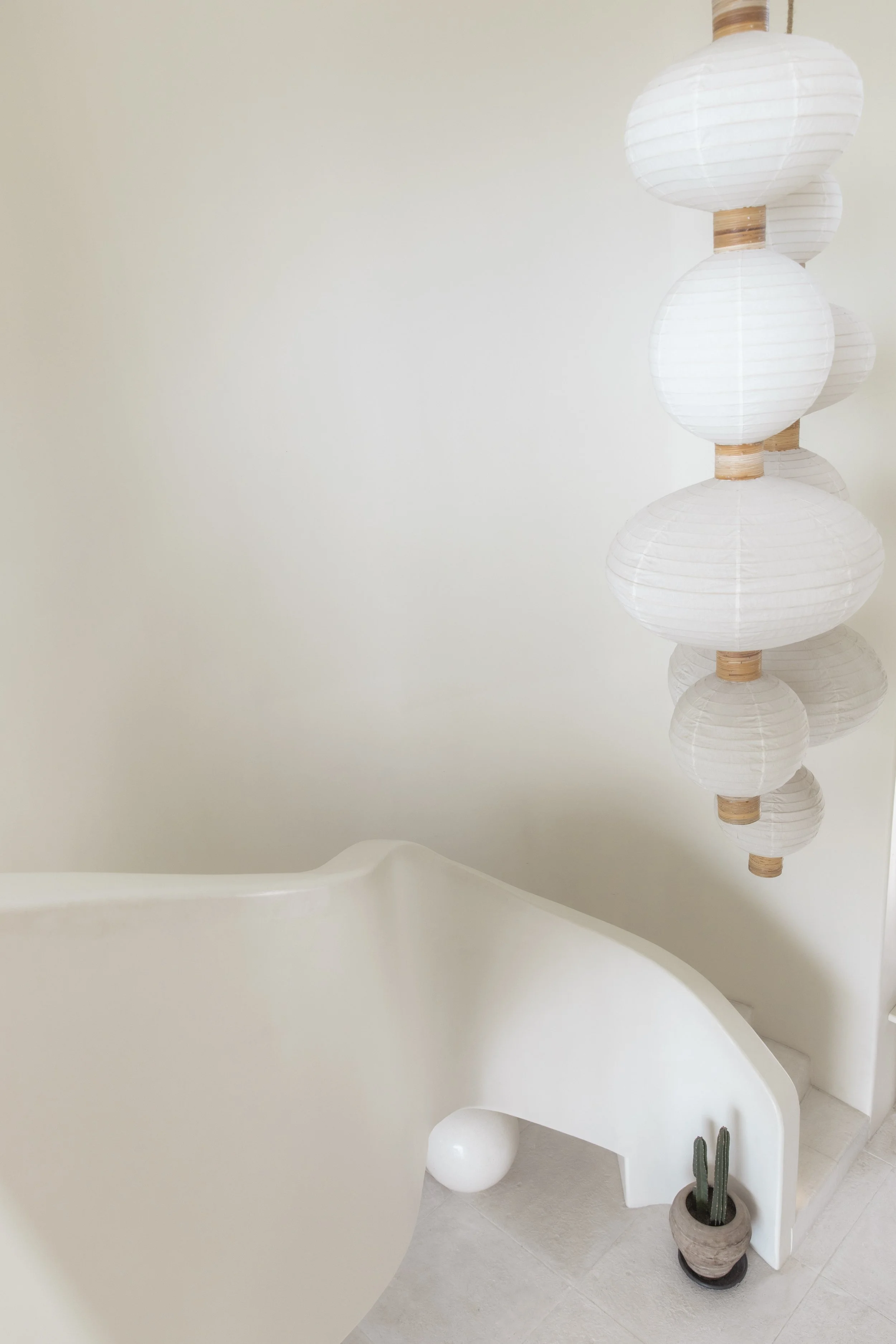 Minimalist white interior with a sculptural curved staircase, clustered paper lantern pendants, pale stone flooring, and a small cactus accent.