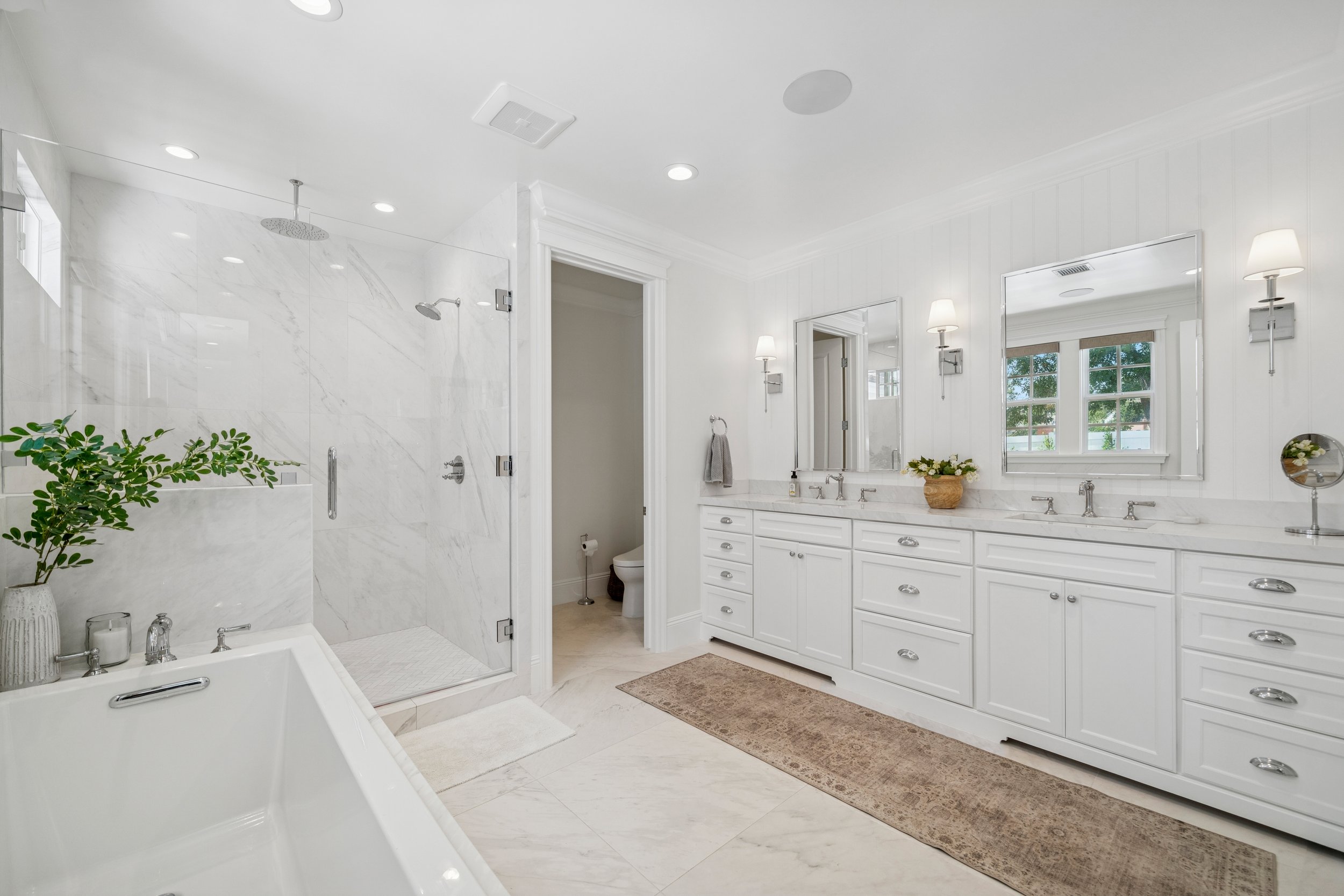 Spa-inspired primary bathroom with white double vanity, marble flooring, glass shower, freestanding tub, chrome fixtures, and bright natural light.