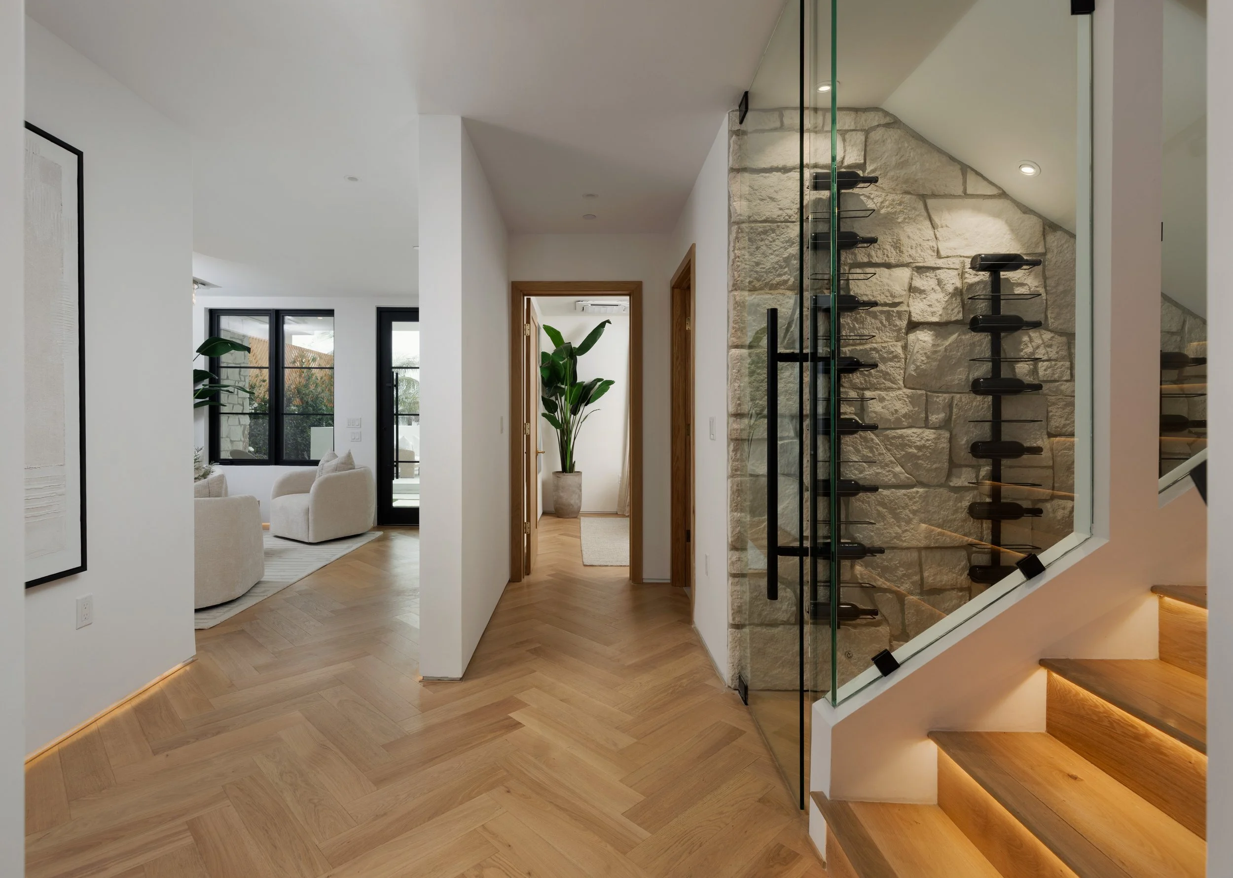 Modern hallway with herringbone wood floors, glass wine storage, stone wall detail, open staircase, and warm stair lighting.