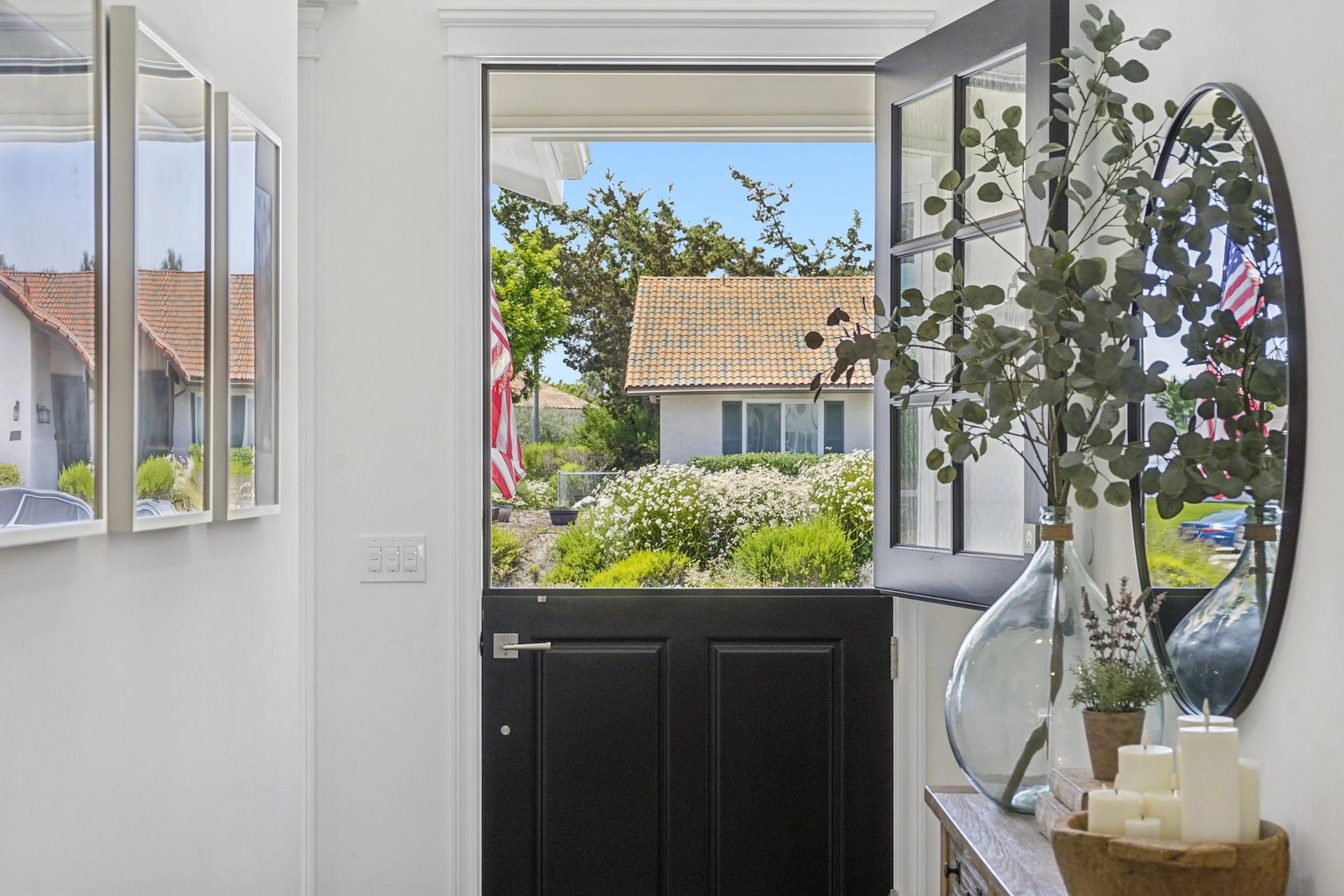 Open black Dutch door in a bright entryway with white walls, round mirror, greenery, and a view of the front landscaping.