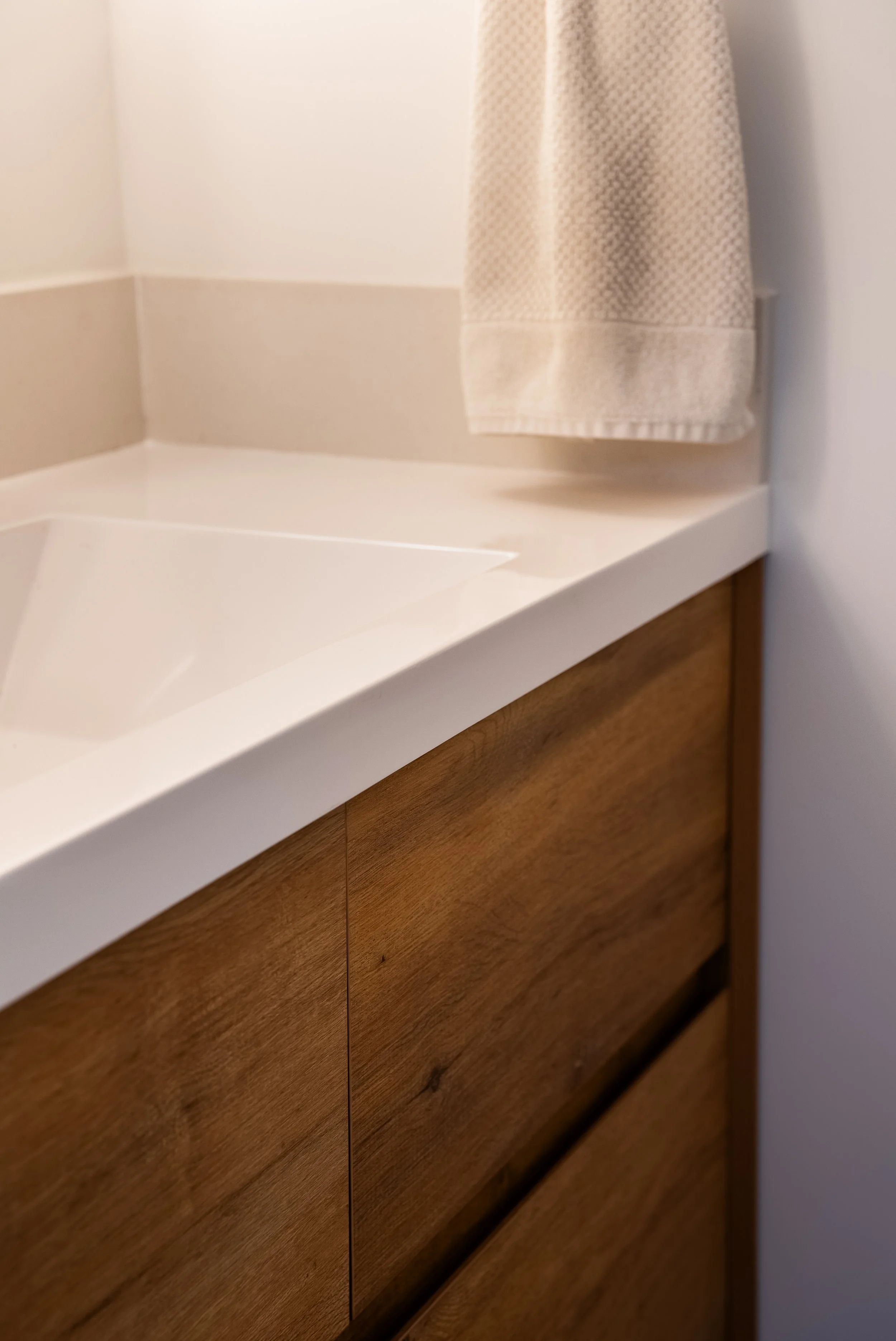 Close-up of a modern ADU bathroom vanity showing wood cabinet detail, white countertop, integrated sink edge, and hanging towel.