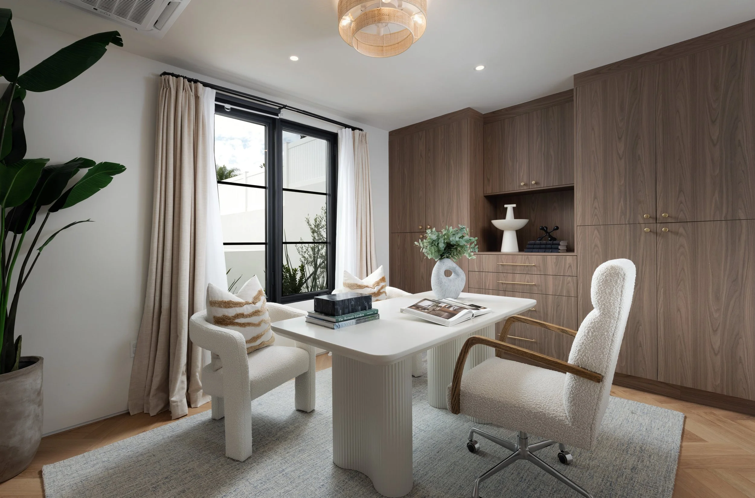 Organic modern home office with walnut built-ins, cream boucle chairs, white desk, black framed windows, soft drapery, and herringbone wood floors.