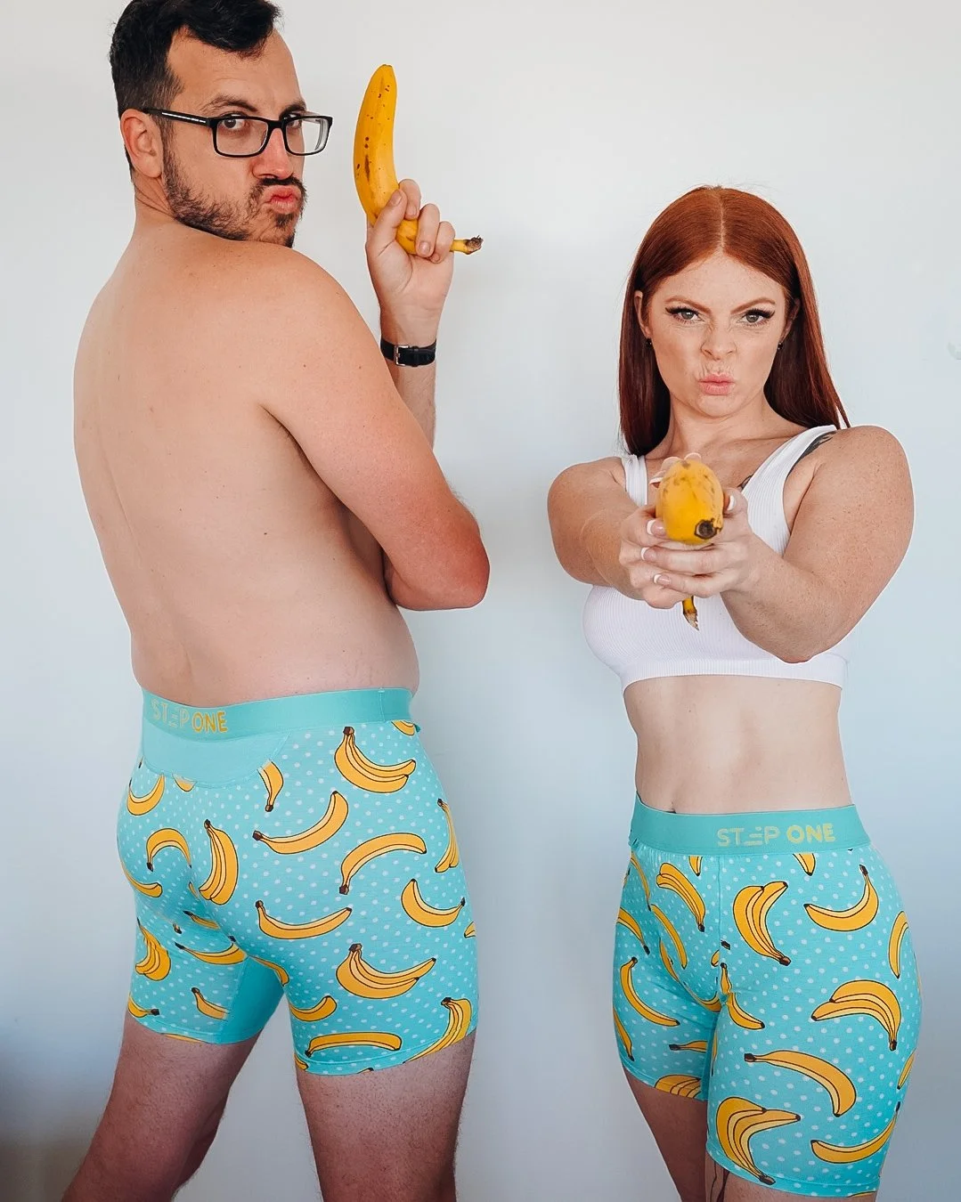 A man and a woman posing with bananas, wearing matching blue underwear with banana print, white Tops for the woman and shirtless for the man, against a plain background.