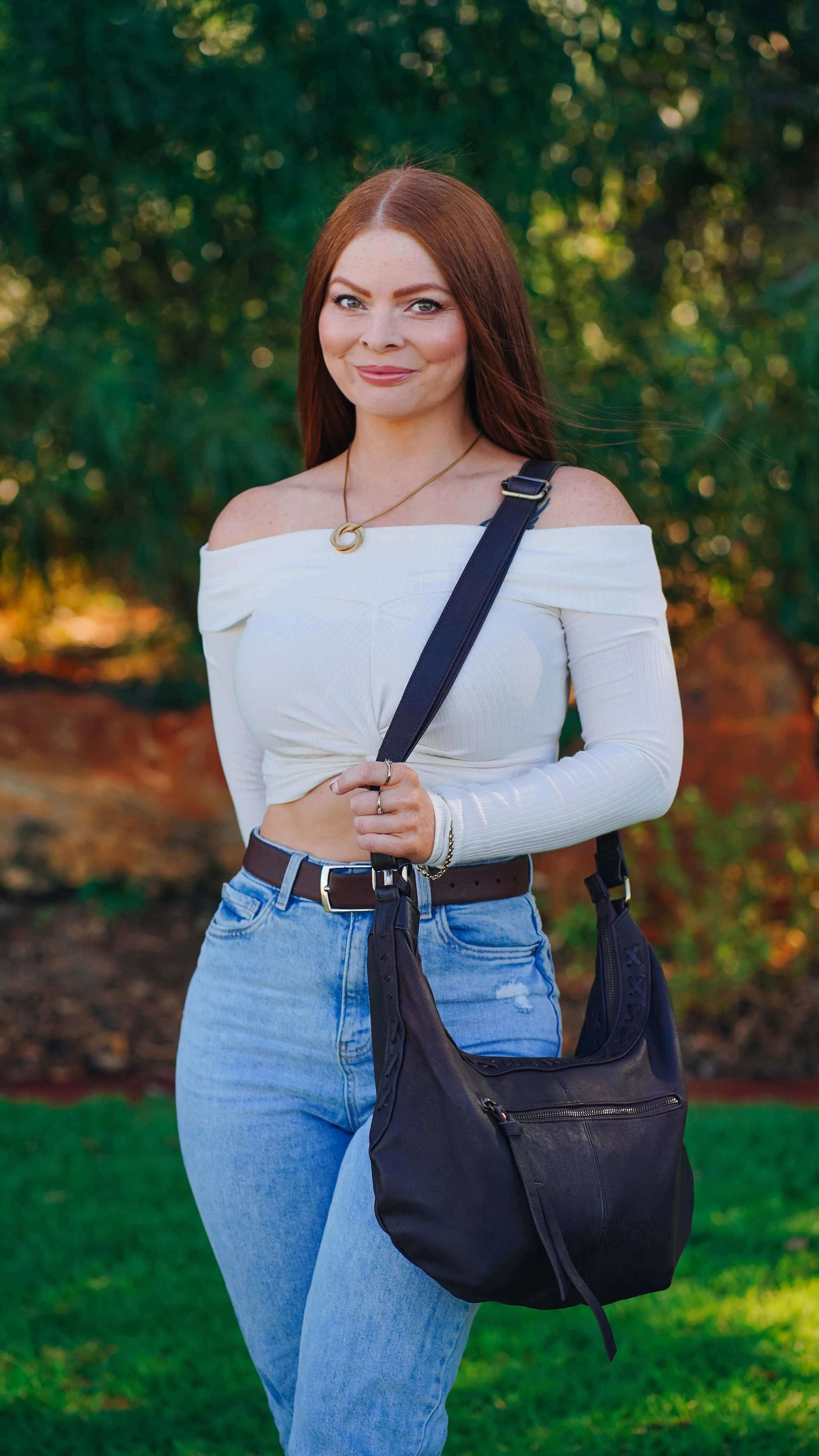 A young woman with long red hair standing outdoors, wearing a white off-the-shoulder crop top, blue jeans, and carrying a black shoulder bag. She is smiling at the camera with greenery in the background.