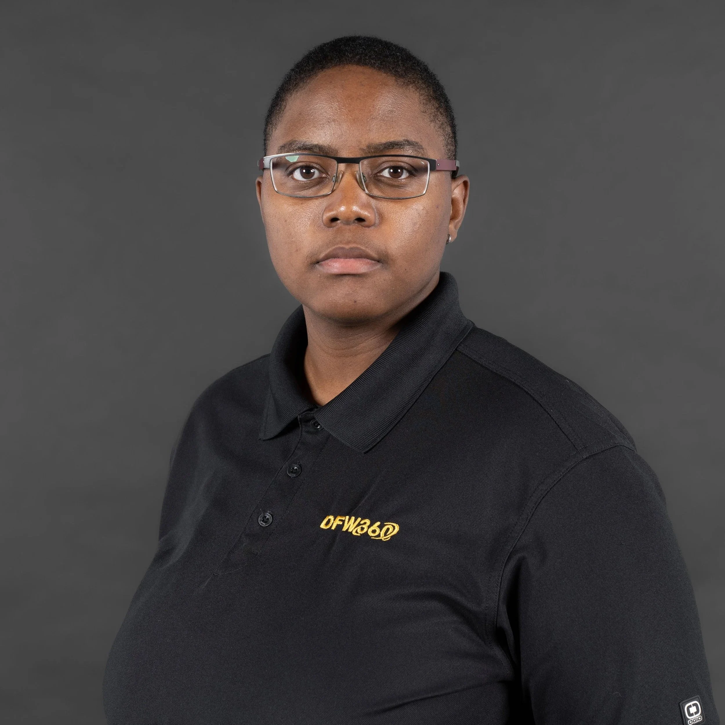 A woman with short hair and glasses wearing a black polo shirt with embroidered text 'DFW36D' on the left chest, standing against a dark gray background.