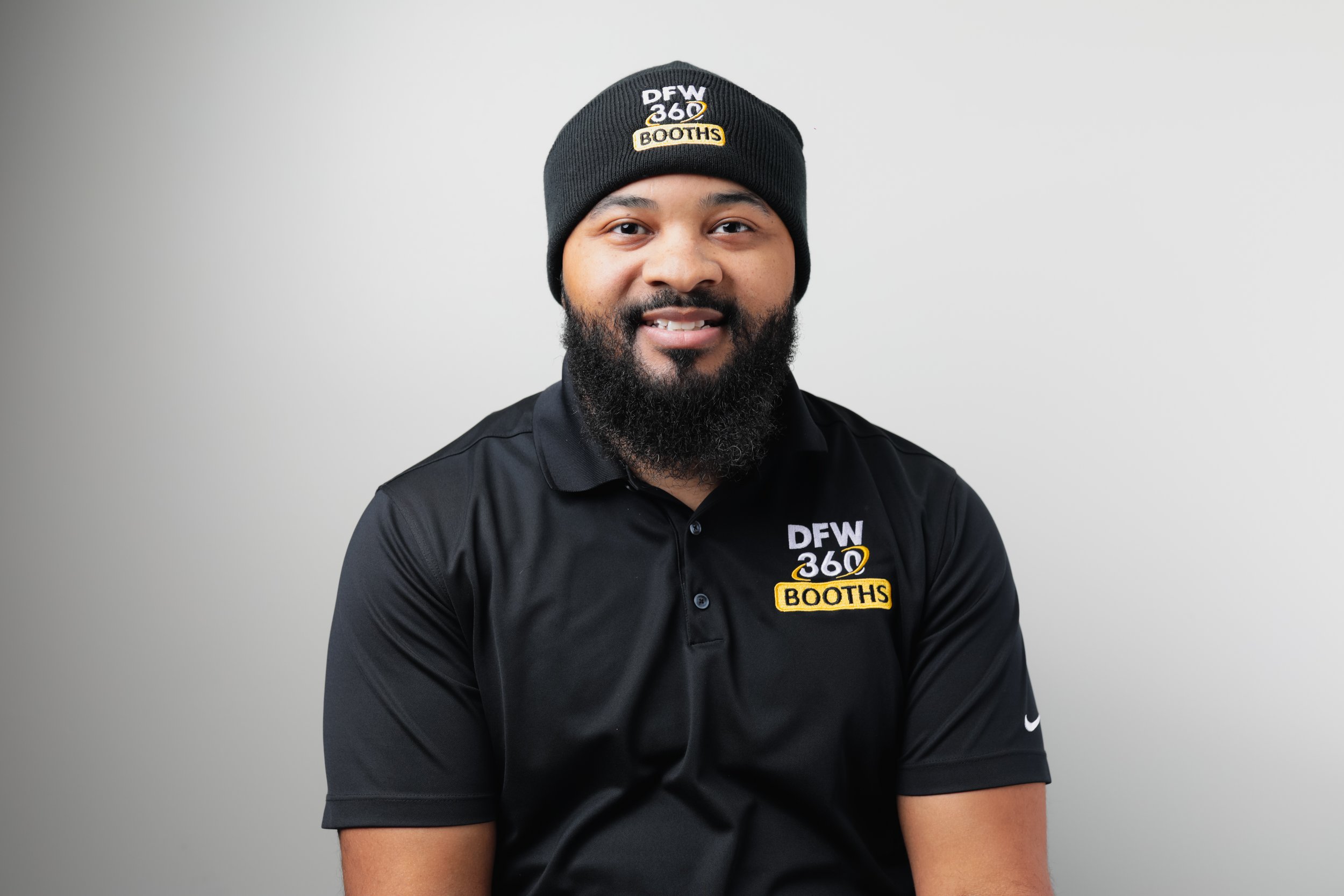 A smiling man with a beard and a shaved head wearing a black polo shirt with a yellow logo and standing against a gray background.