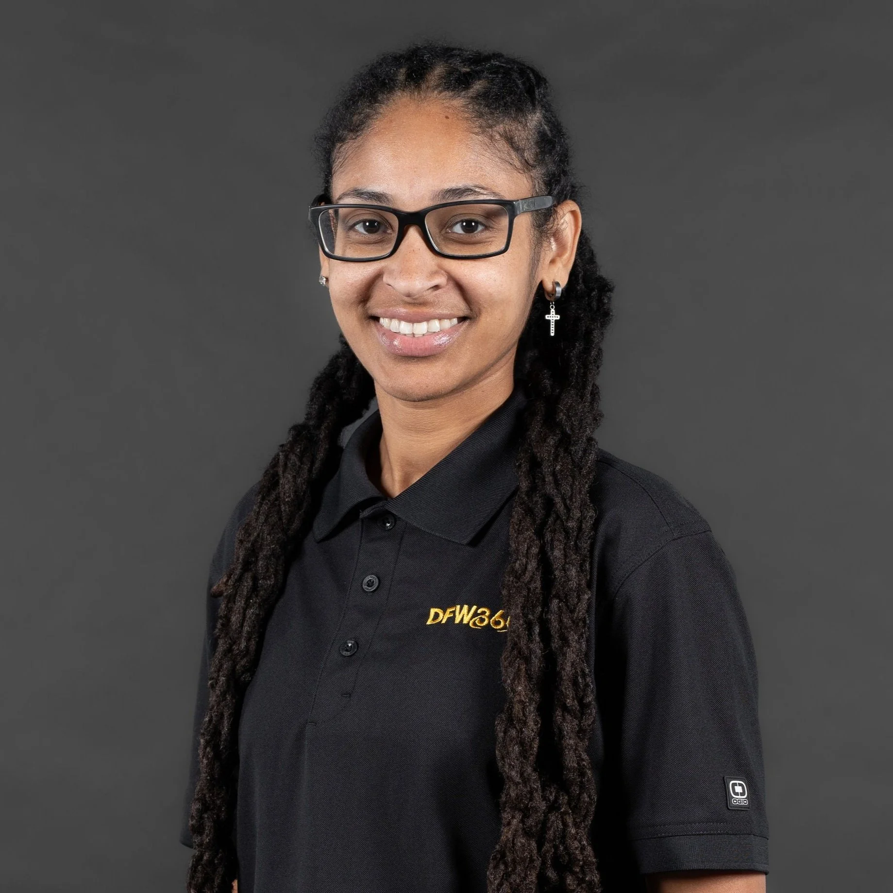 A woman with glasses and long dreadlocks wearing a black polo shirt with yellow text and black pants, standing against a dark gray background.