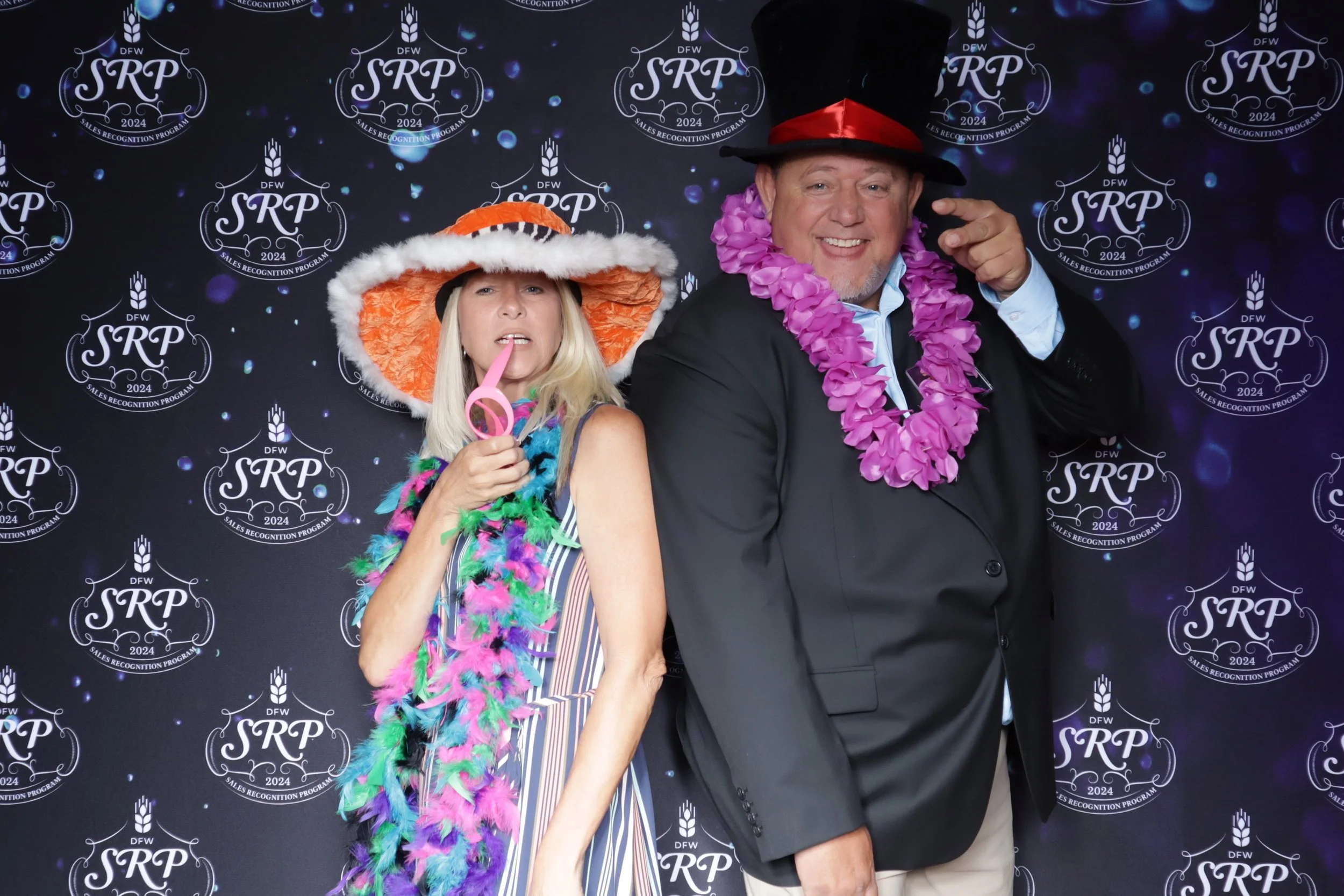 Two people dressed in colorful, playful costumes standing in front of a backdrop with 'DFW SRP 2024' logos. The woman on the left wears a wide-brimmed orange and white hat, a multicolored feather boa, and holds a pink laser pointer. The man on the right wears a black hat with red band, a pink lei, and a black blazer, pointing at the camera.