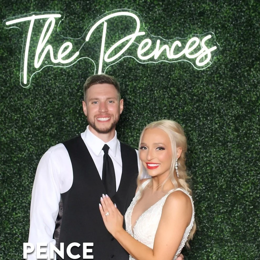 A smiling couple dressed in formal wedding attire standing in front of a green hedge wall with a neon sign that reads 'The Dences.' The man has short brown hair and a beard, wearing a black vest and tie over a white shirt. The woman has long blonde hair, bright red lipstick, and is wearing a white lace wedding dress with earrings, with her hand displaying an engagement ring on her left ring finger.