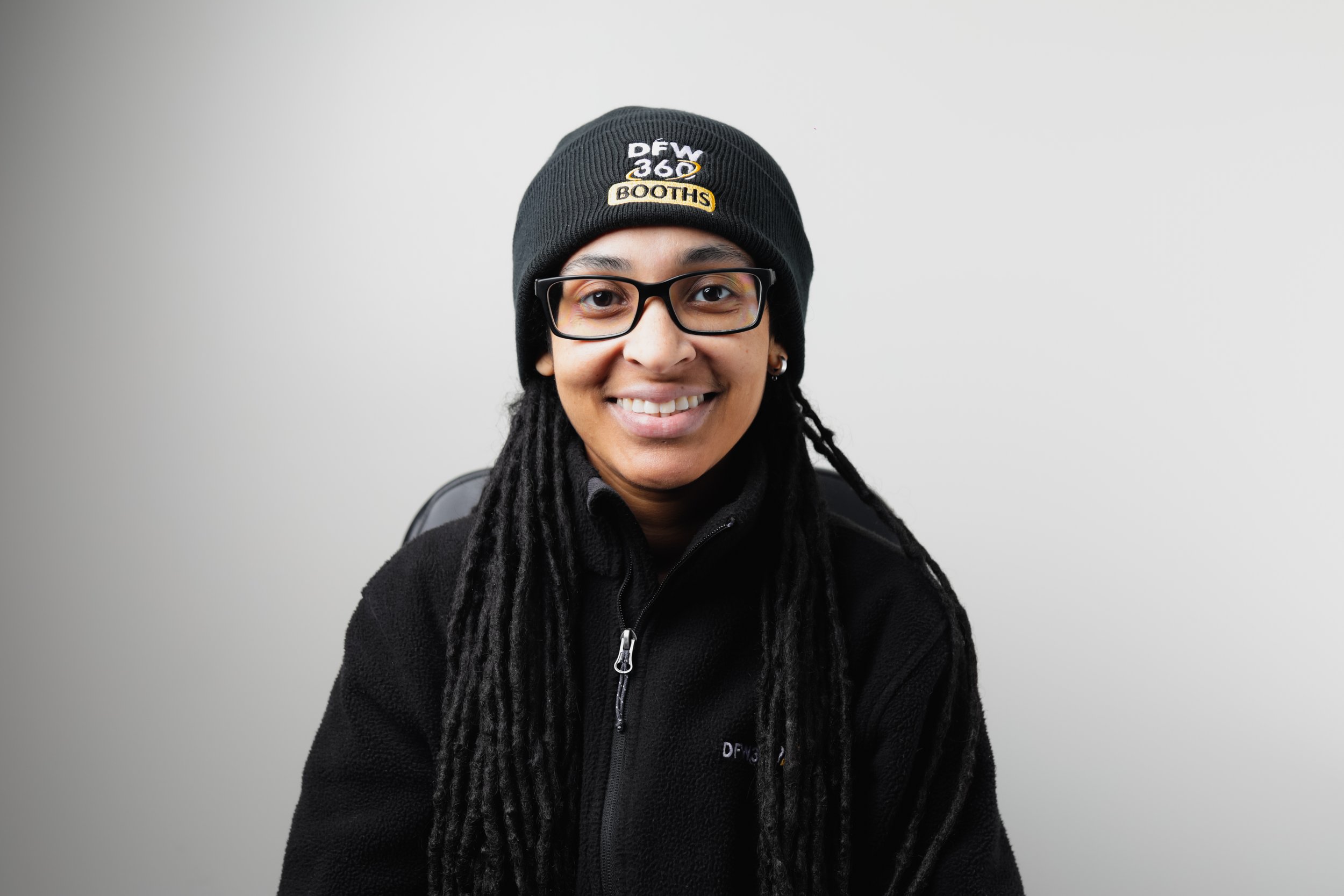 A woman with glasses and long dreadlocks wearing a black polo shirt with yellow text and black pants, standing against a dark gray background.