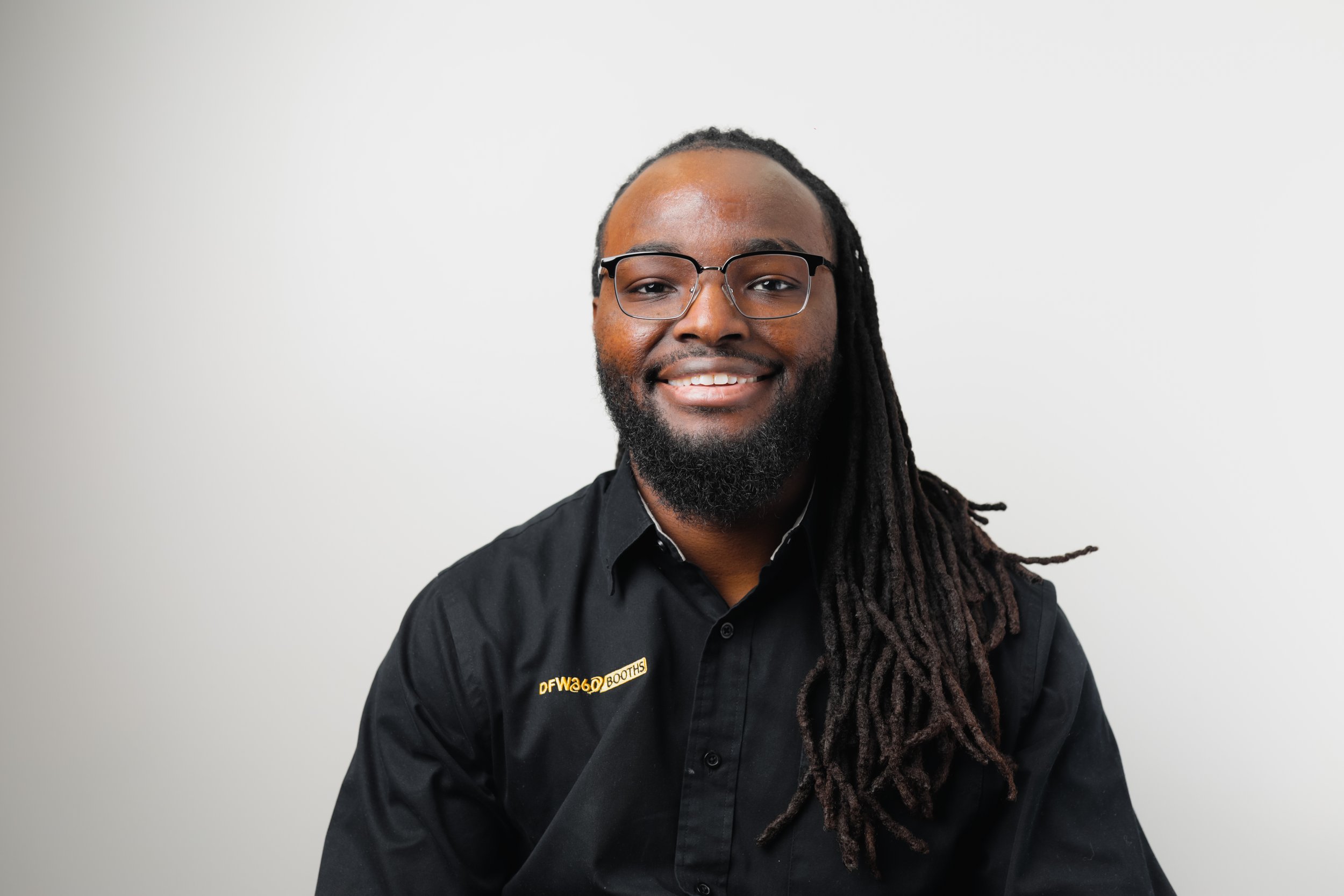 Portrait of a smiling Black man with long dreadlocks and a beard, wearing a black collared shirt with a logo that says DFW360.