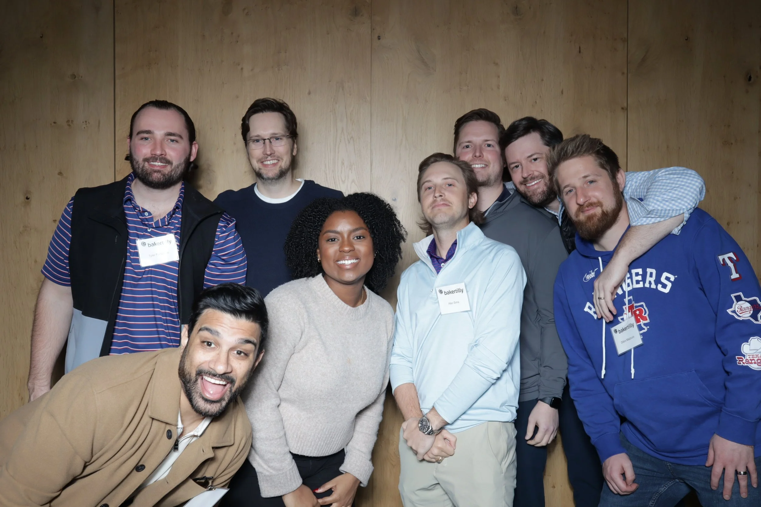 Group of nine diverse people smiling and posing together indoors with a wooden wall background, some wearing name tags.