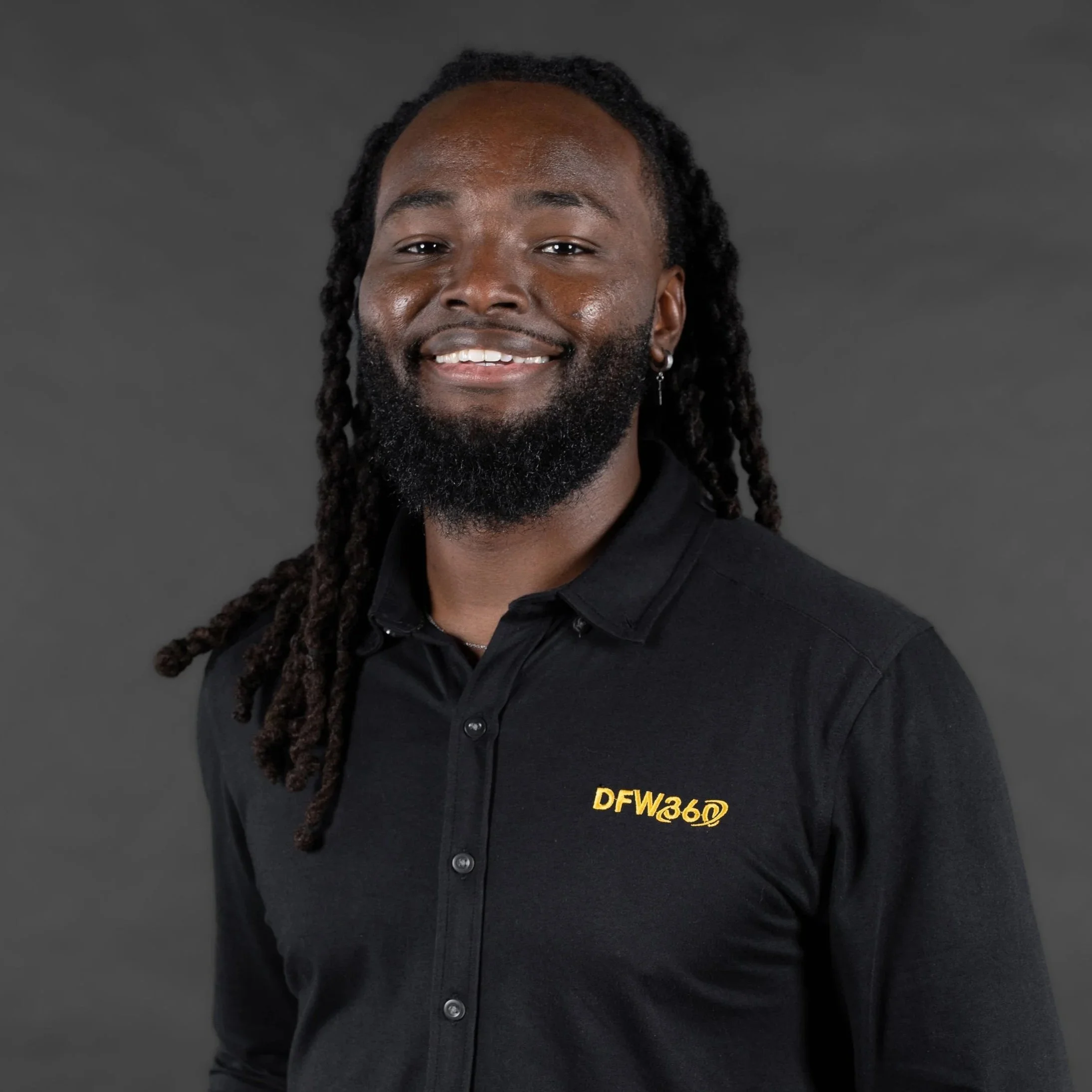 Portrait of a smiling Black man with long dreadlocks and a beard, wearing a black collared shirt with a logo that says DFW360.