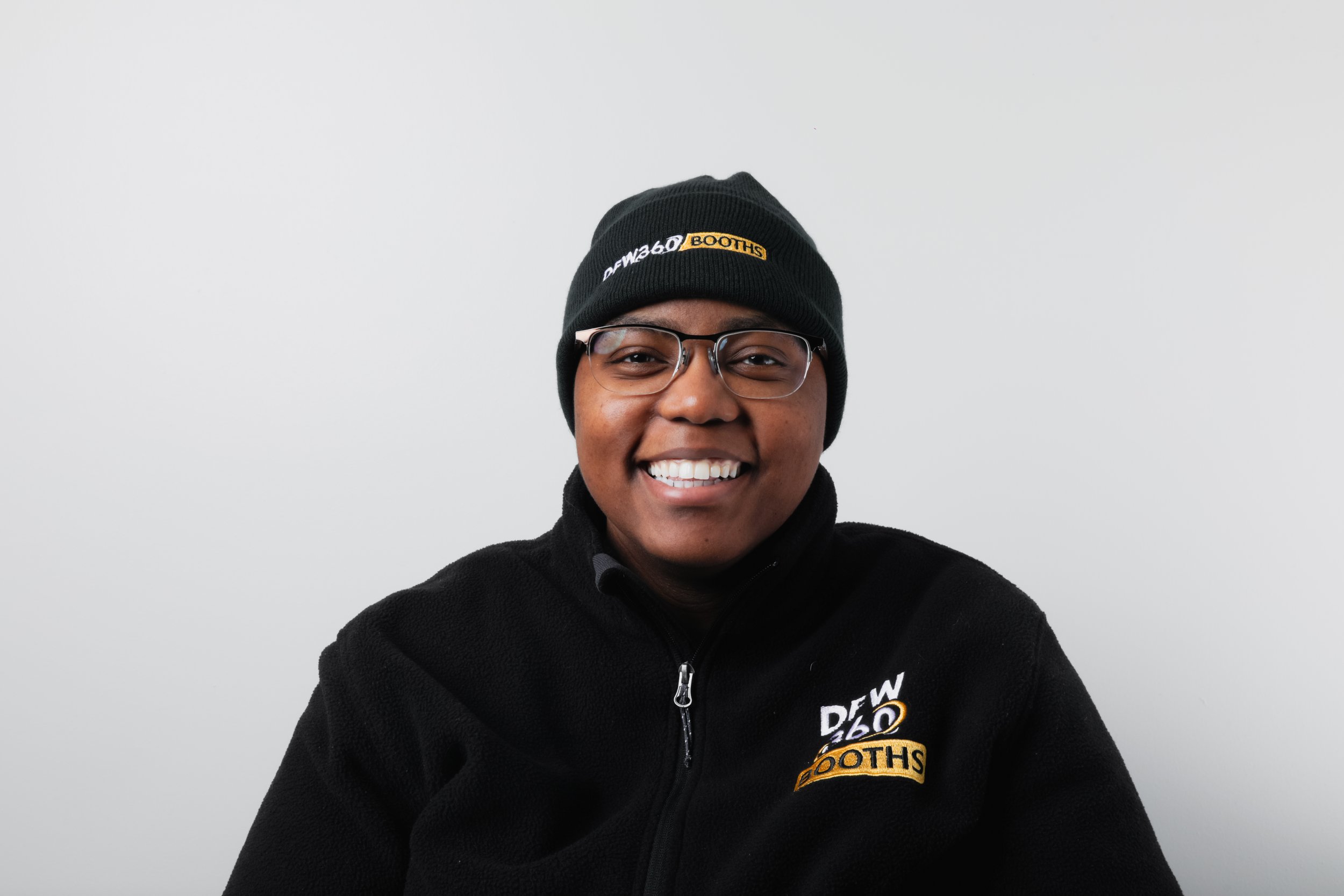 A woman with short hair and glasses wearing a black polo shirt with embroidered text 'DFW36D' on the left chest, standing against a dark gray background.