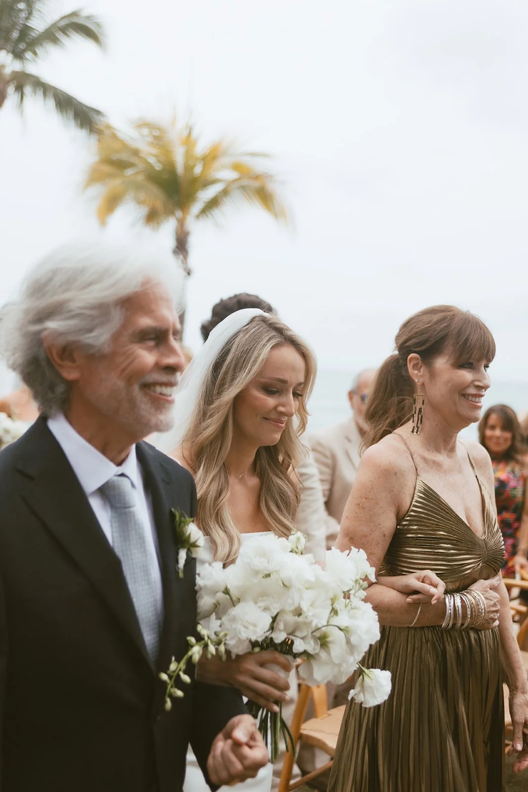 Bride walking down the aisle with her parents, carrying an all-white bouquet