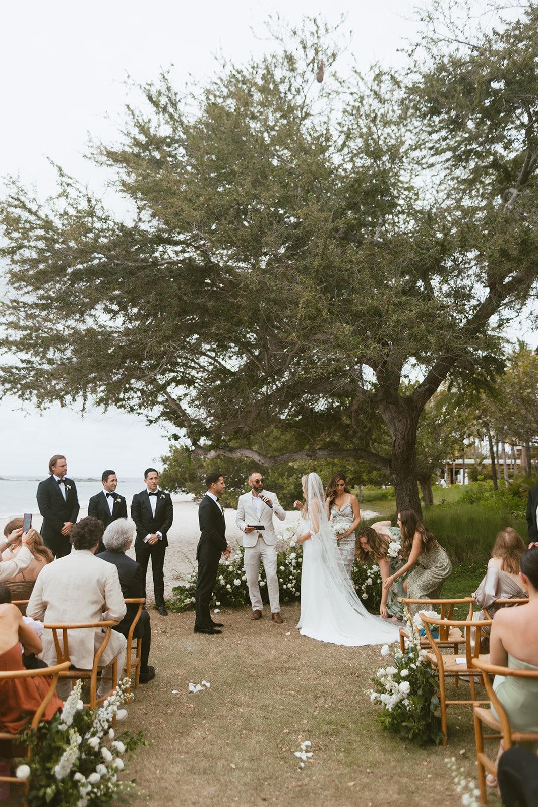 Wedding ceremony under the tree with the bay behind, white ground florals framing the couple
