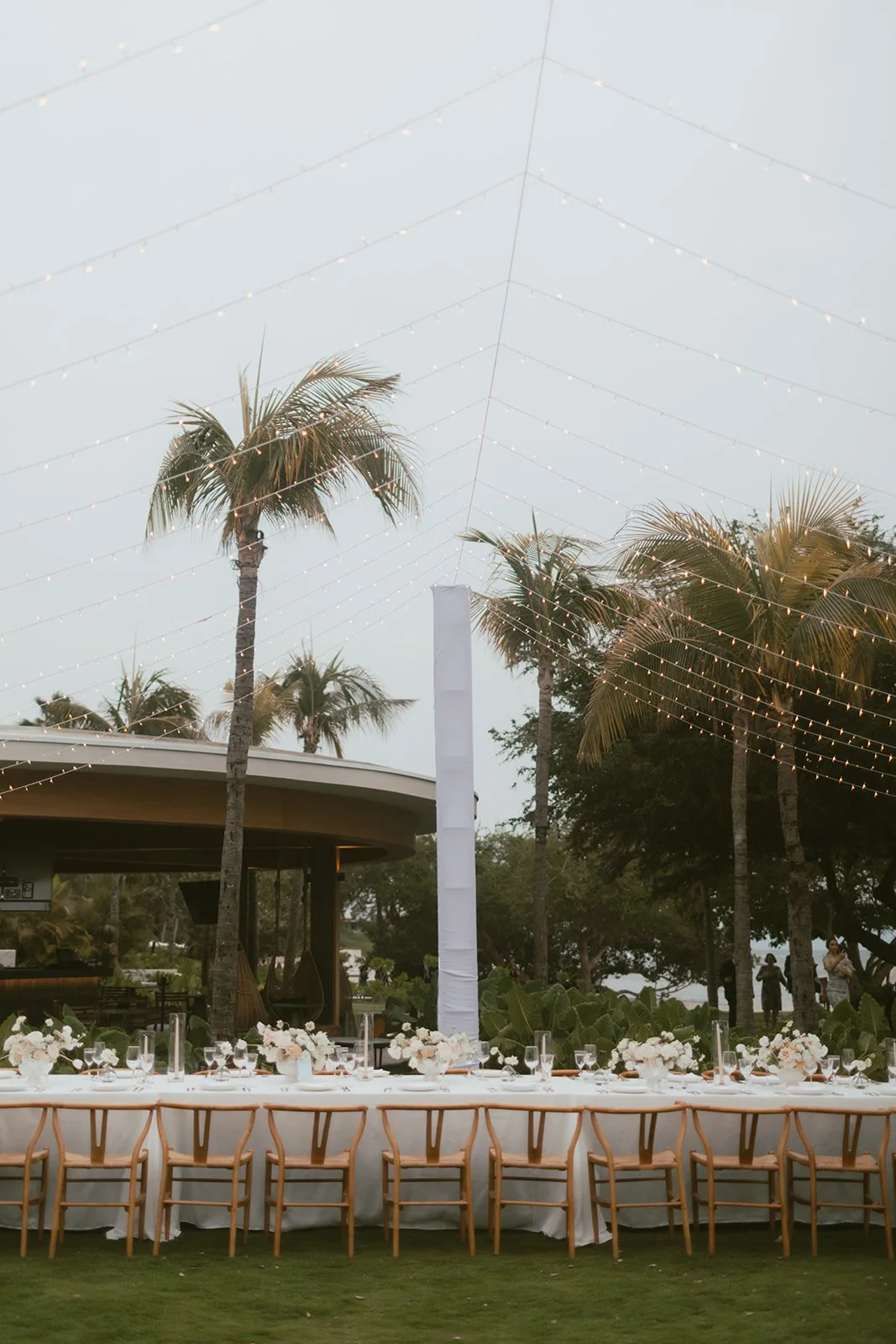Long reception table under string lights on the lawn, white and green centerpieces
