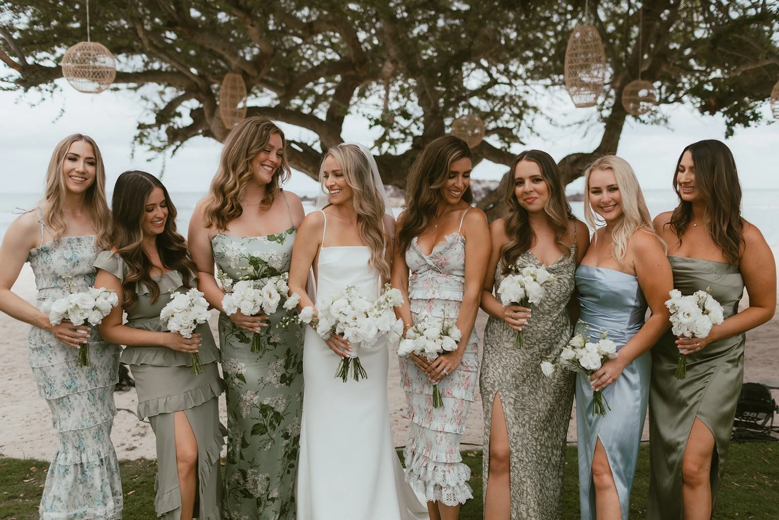 Bridesmaids in sage and blue holding all-white bouquets on the beach lawn