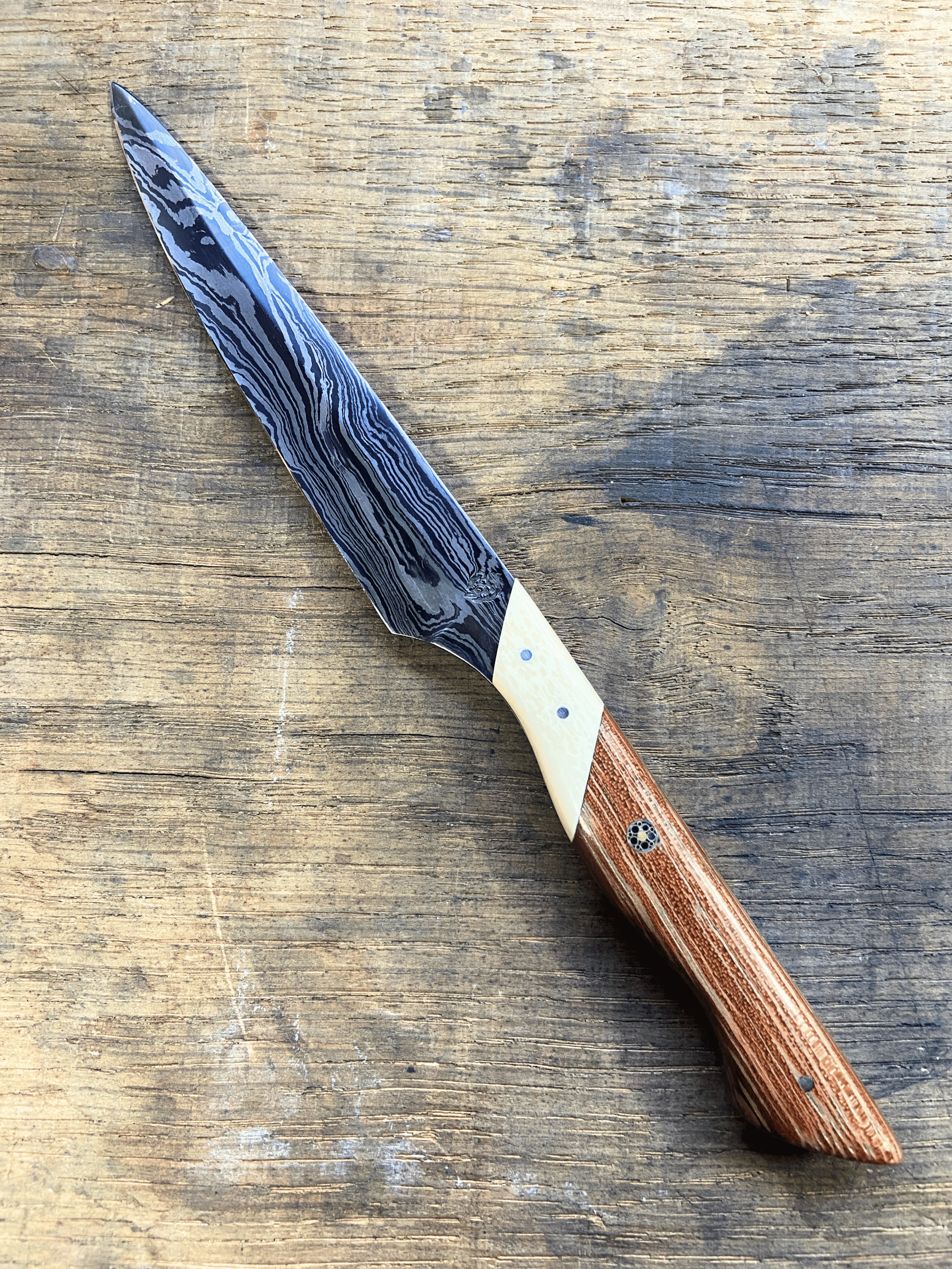 Un couteau en acier damassé noir et argenté, avec manche blanc et brun clair sur fon de bois brun.