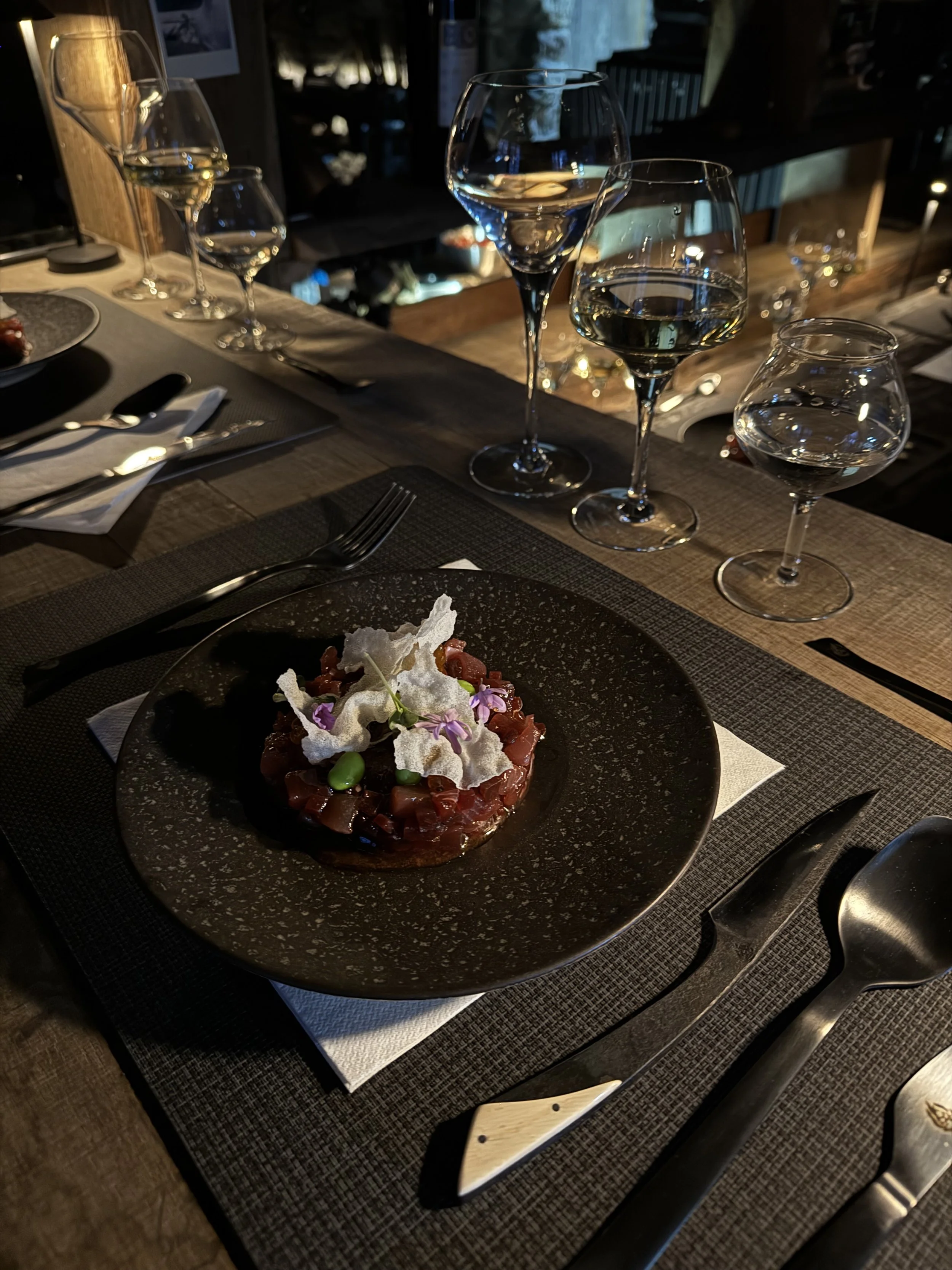 Une assiette contenant un tartare de poisson, décoré de fleurs, de croustilles de parmesan, sur une table en bois avec des plats et des verres à vin.