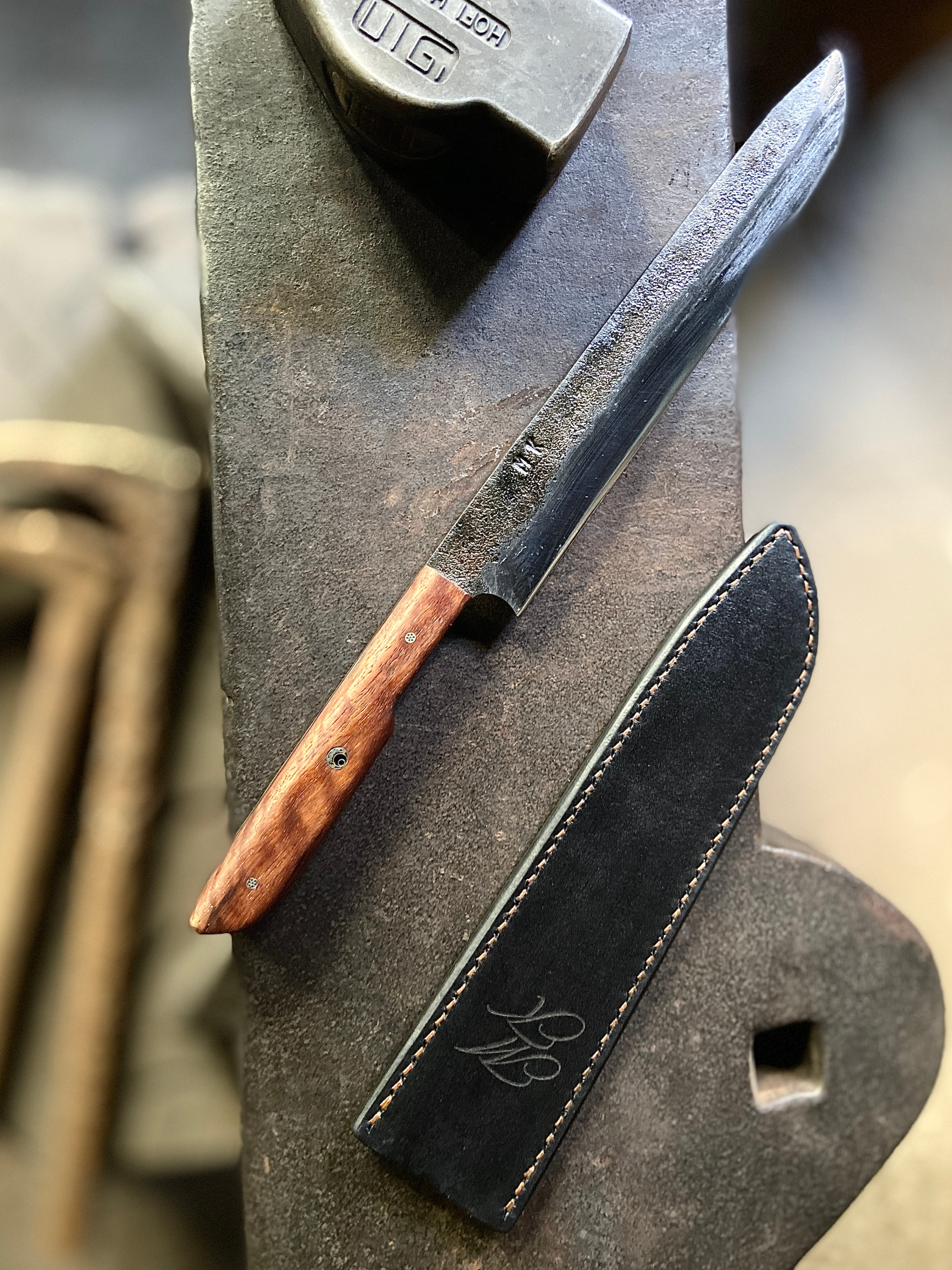 Couteau de Chef forgé brut de forge avec manche en bois marron à côté de son étui en cuir foncé et à côté d'un marteau de forgeron sur fond d'enclume grise.