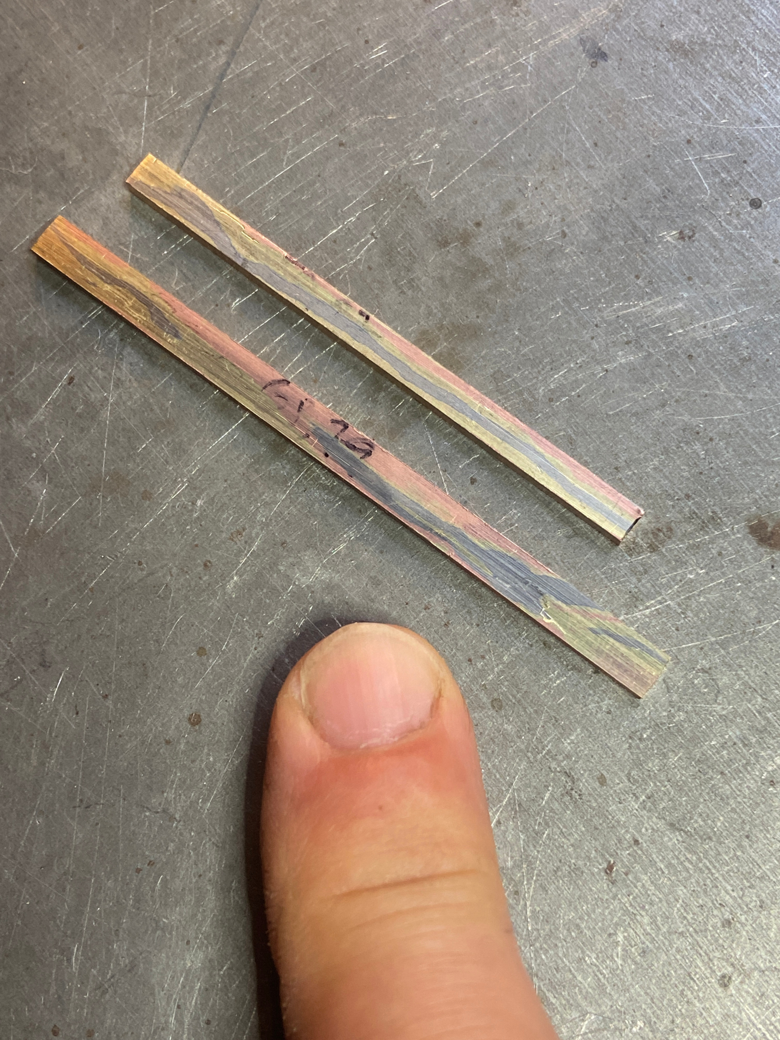 Une personne montre deux petits morceaux de métal imitation bois, avec un doigt visible au premier plan sur une surface métallique.
