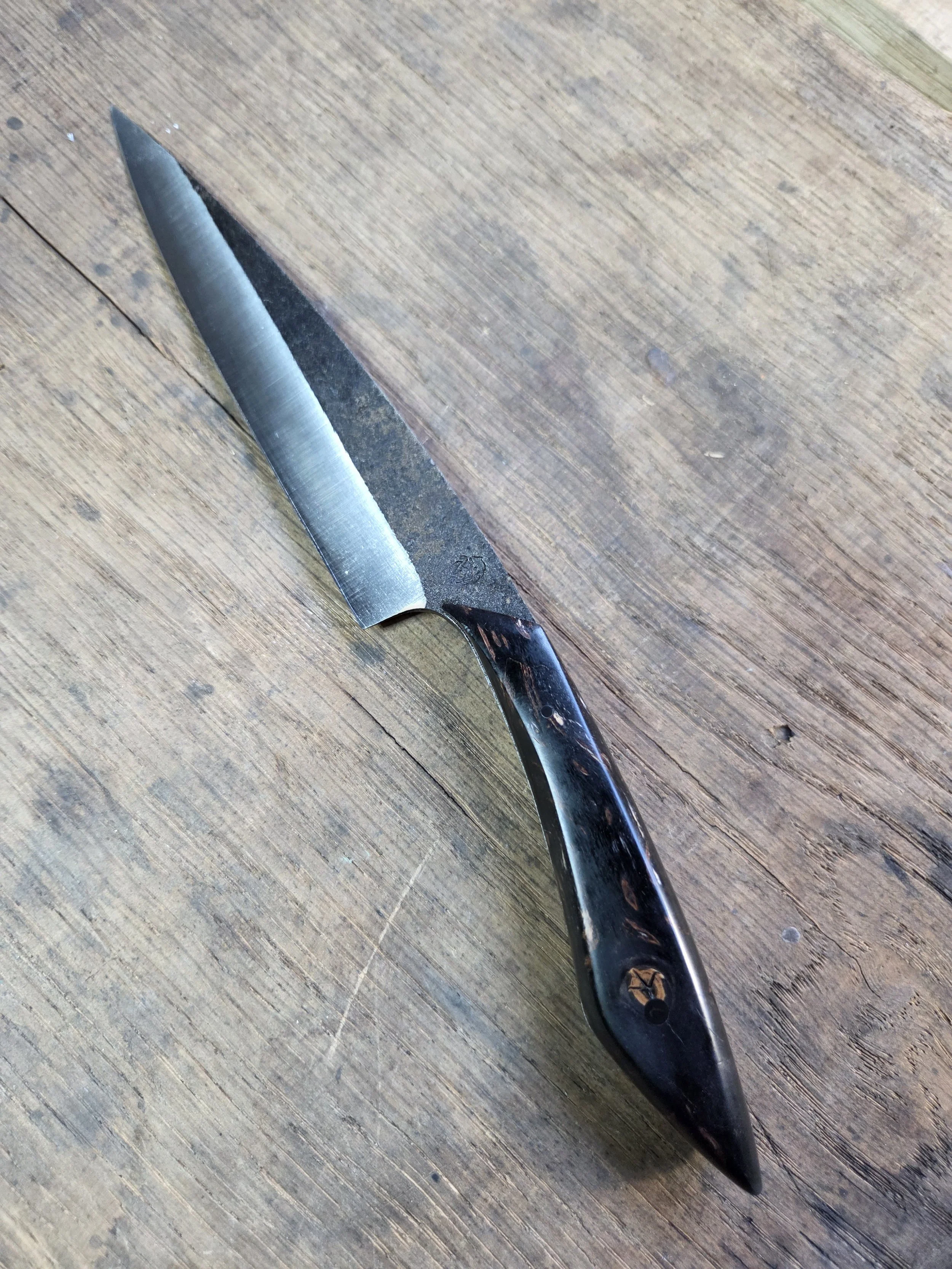 Couteau forgé brut de forge avec manche en bois noir et tâches oranges sur fond de bois de chêne marron.