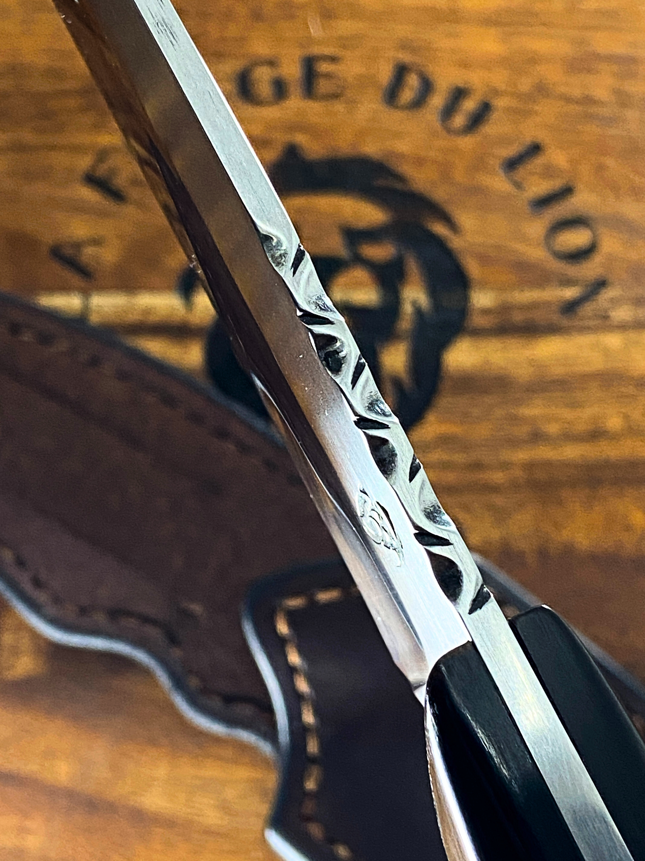 Couteau droit forgé avec une décoration gravée sur le dos du couteau, au-dessus d'un étui en cuir brun posé sur un fond en bois brun  avec un logo de tête de lion et une inscription LA FORGE DU LION.