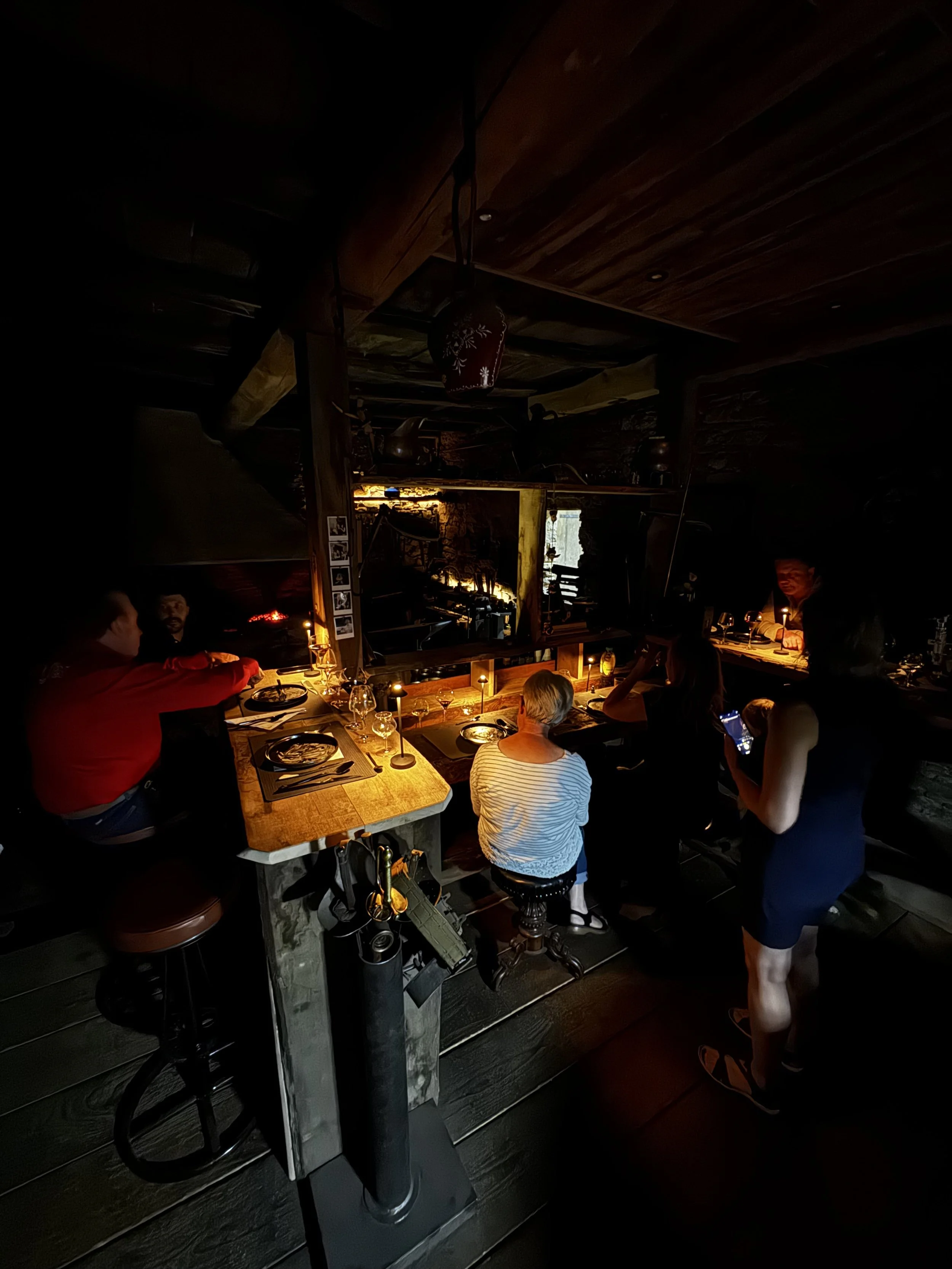 Une scène à l'intérieur d'un restaurant ou d'un bar en basse lumière, avec plusieurs personnes assises et debout autour d'une table en bois. La lumière est principalement concentrée sur la table, créant une ambiance chaleureuse. Une femme en robe noire se tient à côté de la table, et une autre personne avec des cheveux blonds et en chemise rayée est assise, regardant vers la table. Un décor en bois et quelques affiches ou cadres sont visibles sur le mur.